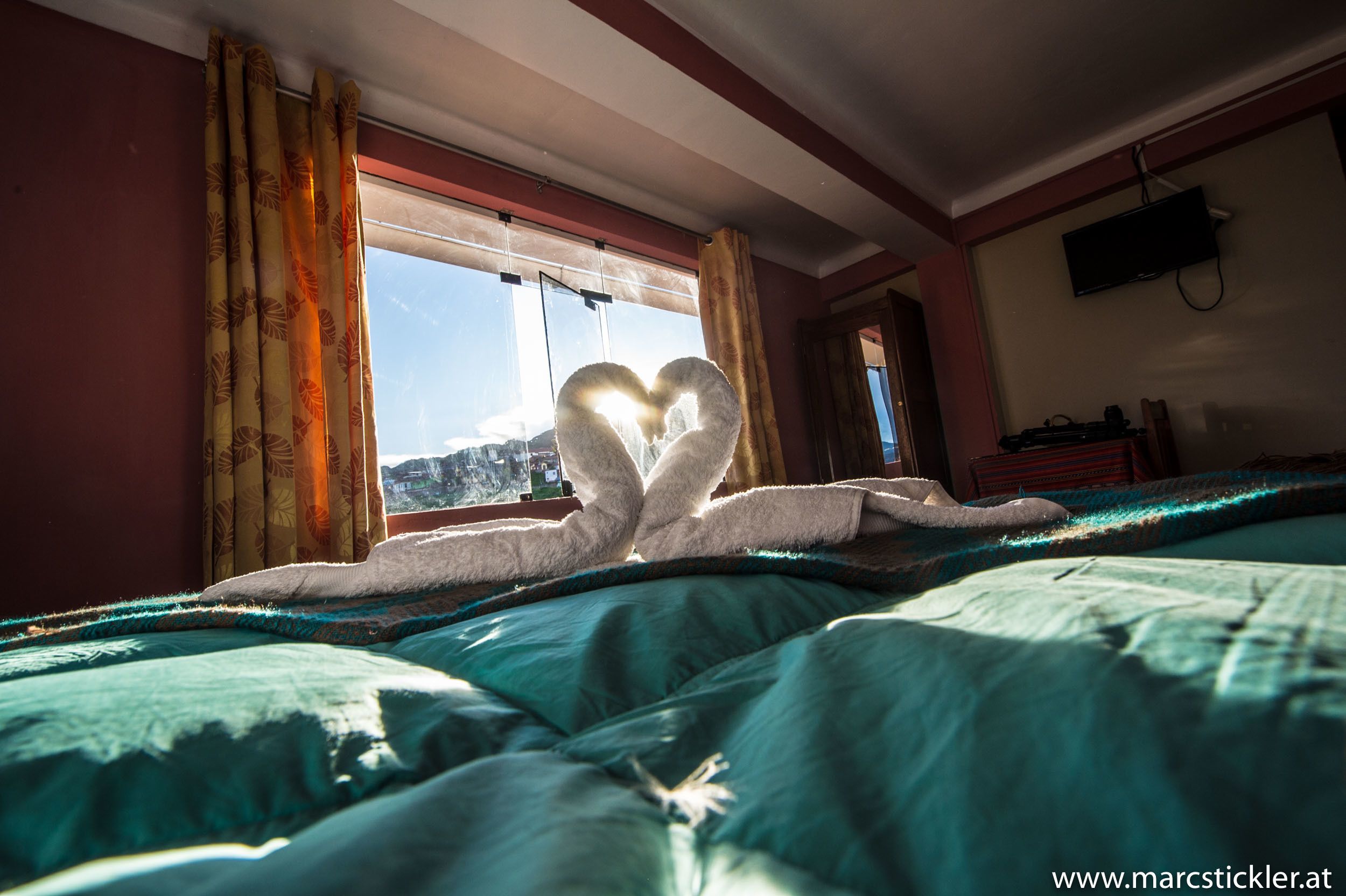 A hotel room with a bed, a window with curtains, a flat-screen TV on the wall, and a view of a mountain.