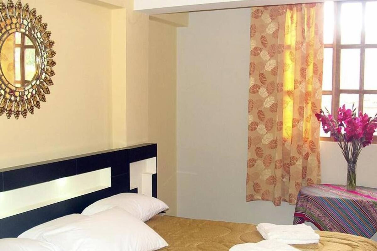 A bed with white pillows and a quilt, a wall-mounted mirror, a window with floral curtains, and a vase with flowers.