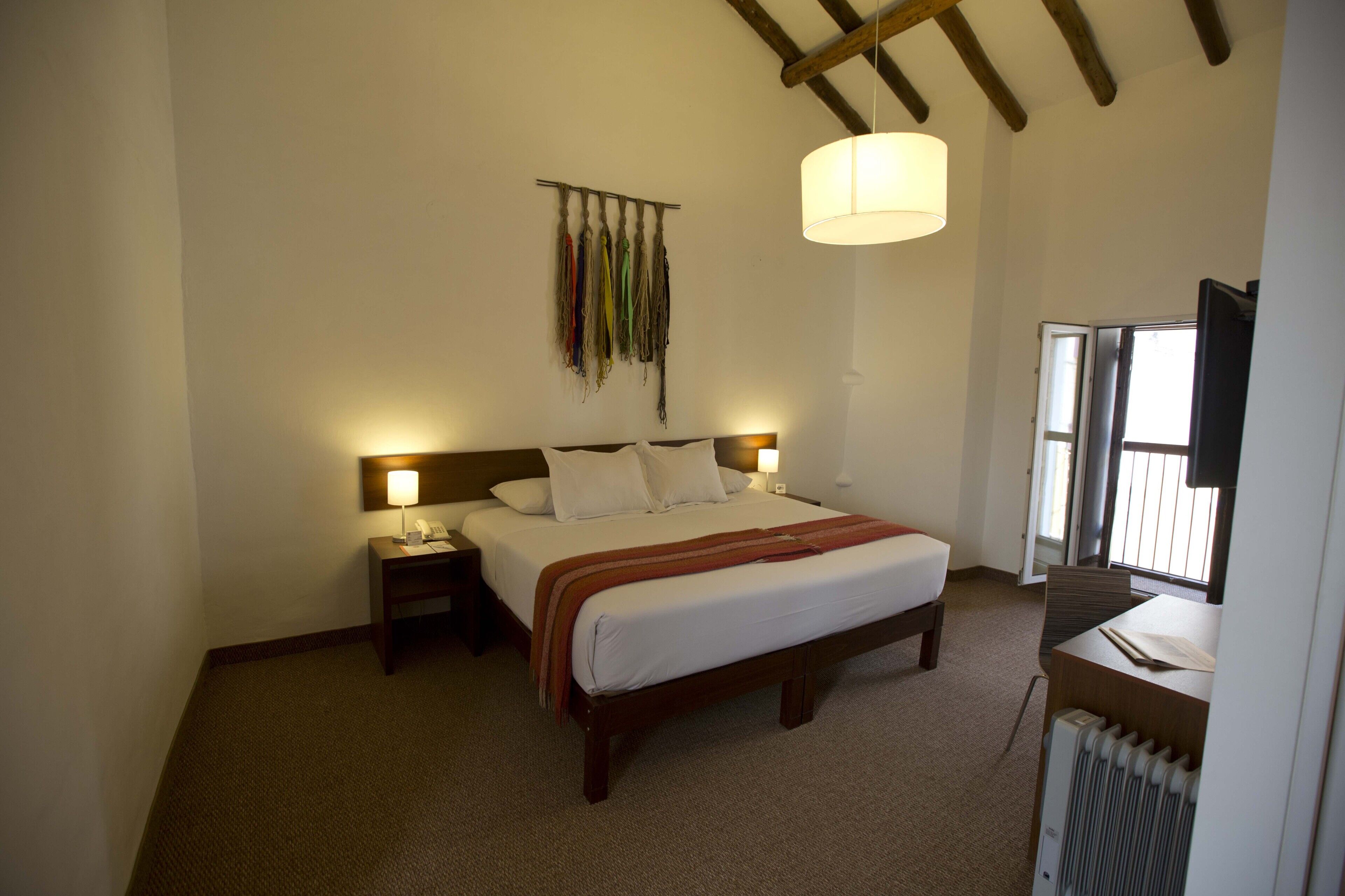 A hotel room with a bed, bedside tables, a lamp, a radiator, and a window with blinds.