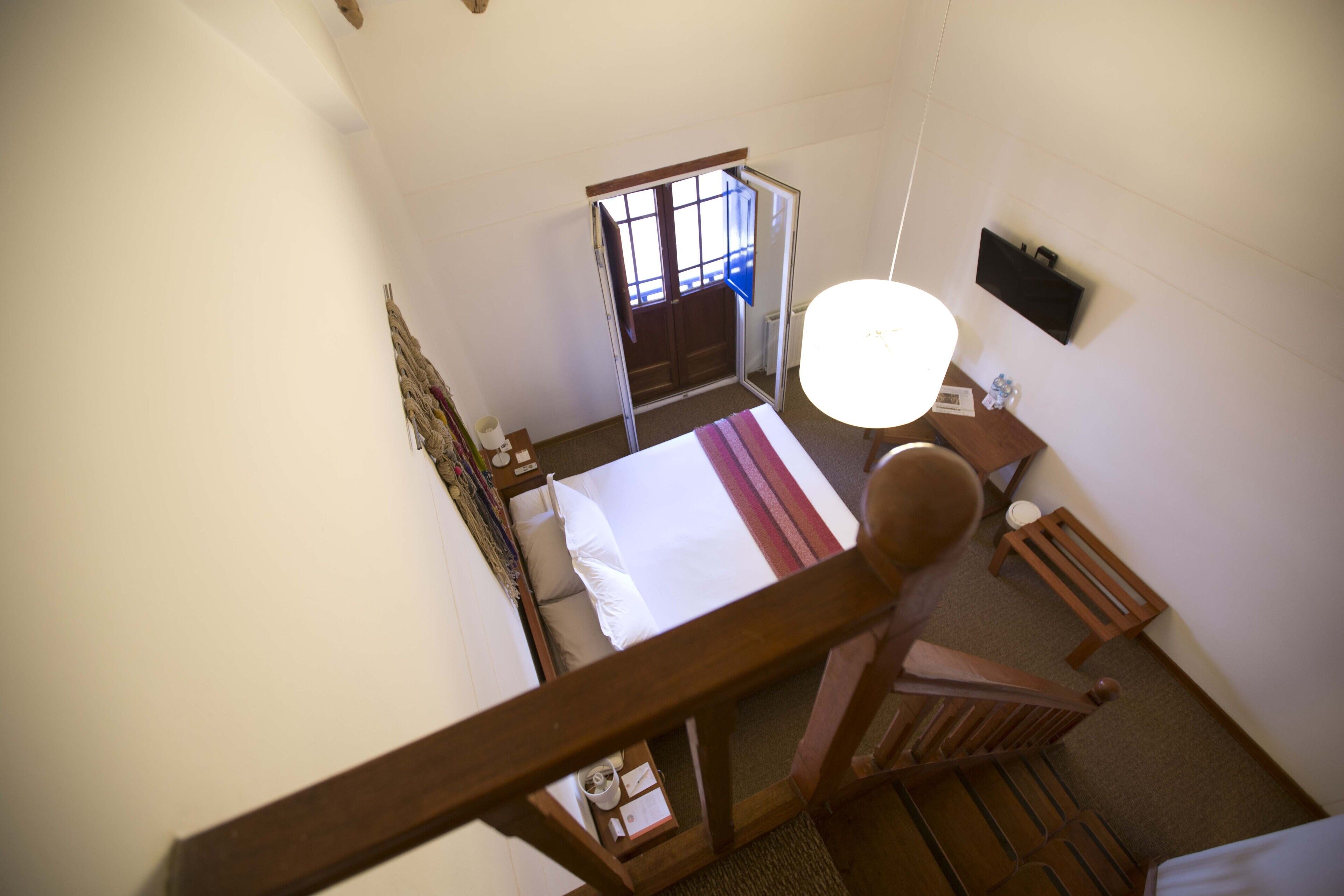 A hotel room with a bed, a wooden chair, a desk with a lamp, and a window with a view of the outside.