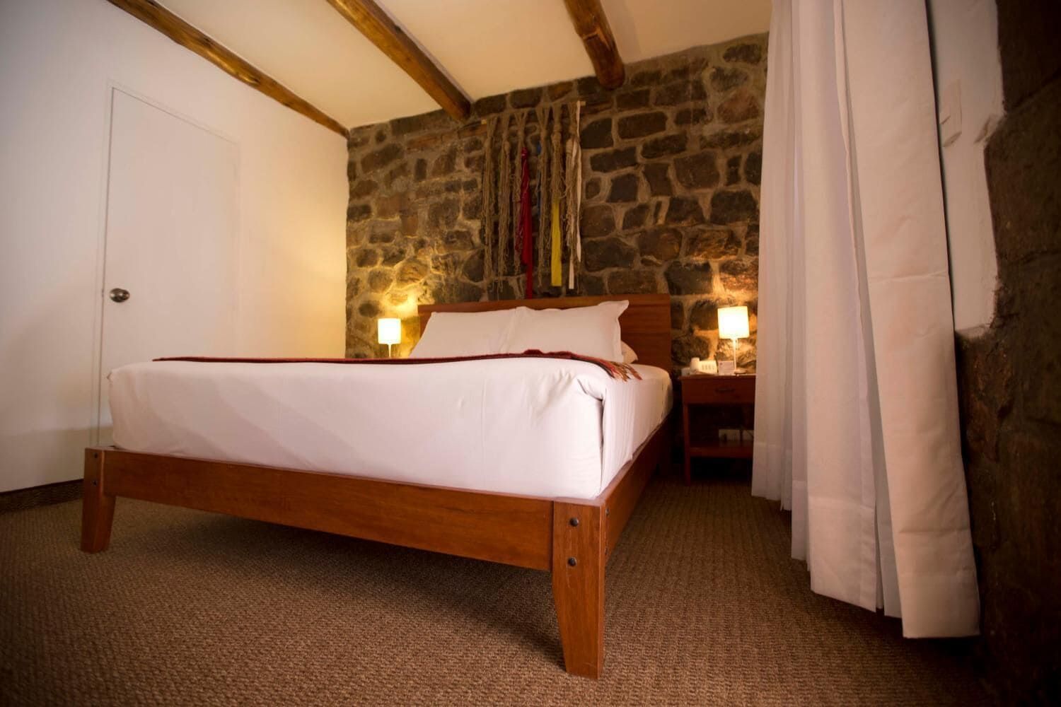 A bedroom with a wooden bed, stone wall, and two bedside tables with lamps.
