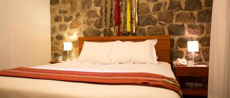 Una cama con sábanas blancas y una manta a rayas rojas y anaranjadas, un cabecero de madera y dos mesitas de noche con lámparas.