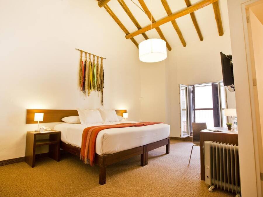 A hotel room with a wooden bed, a nightstand, a desk with a chair, a radiator, and a window with a view.