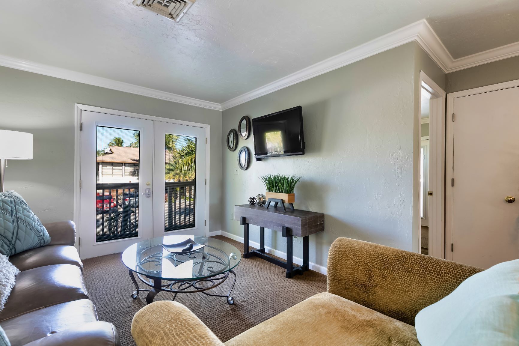 A living room with a glass coffee table, two sofas, a TV mounted on the wall, and a sliding glass door leading to a balcony.