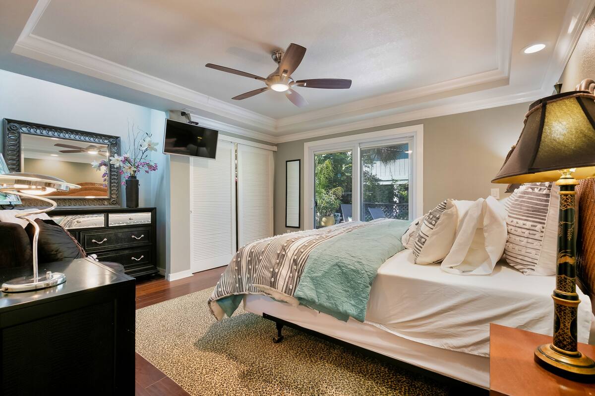 A bedroom with a bed, a ceiling fan, a TV, and a window.