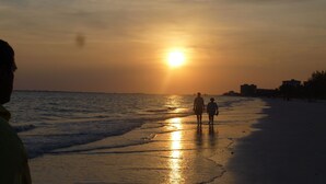 Ein Strandbild mit Sonnenuntergang, Menschen, die am Ufer entlanggehen, und Gebäuden im Hintergrund.