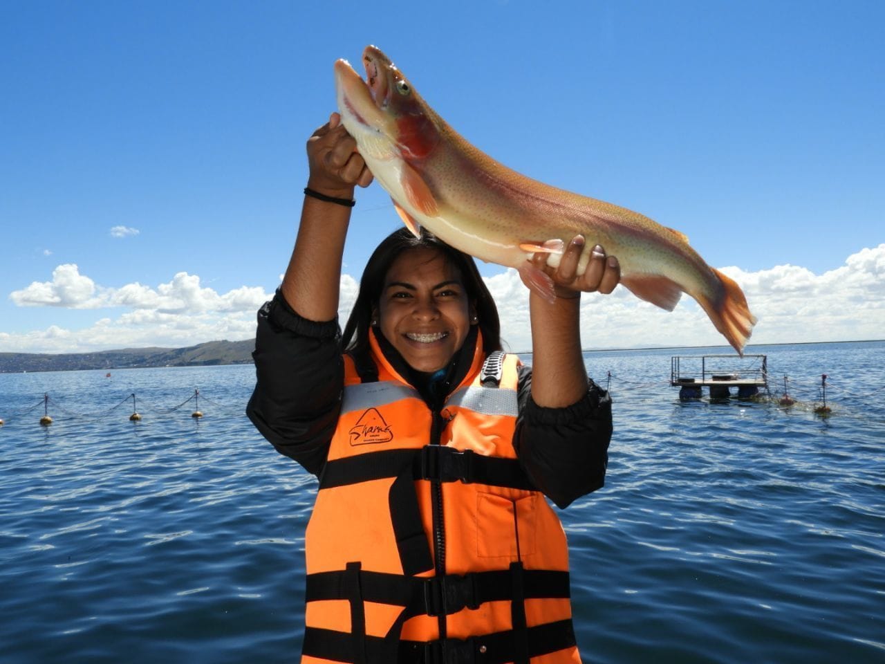 Una persona sosteniendo un pez por encima de la cabeza, con chaleco salvavidas, y un cuerpo de agua y un bote al fondo.