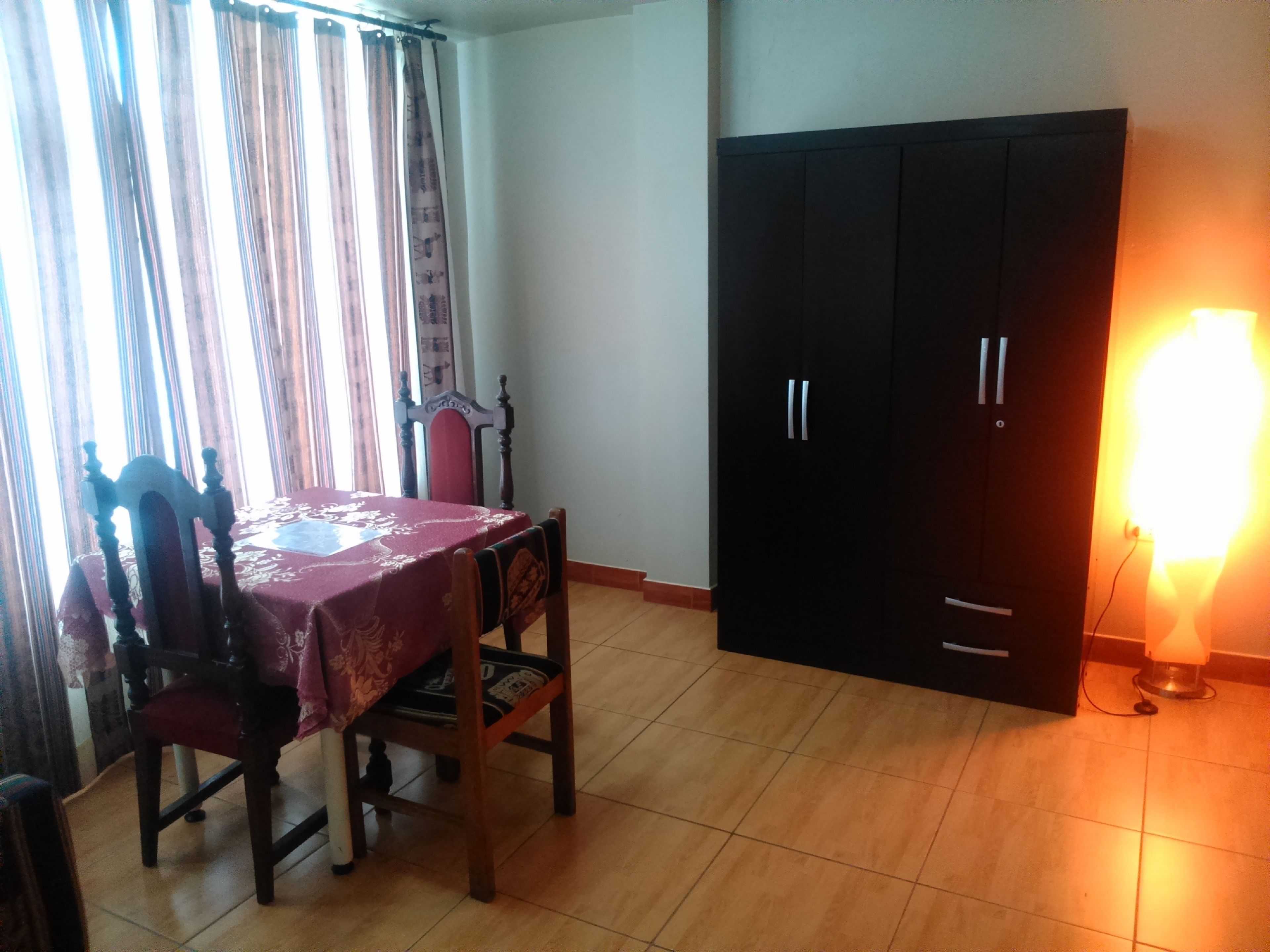 A room with a dining table covered with a red tablecloth, two wooden chairs, a black wardrobe, and a bright lamp on the floor.