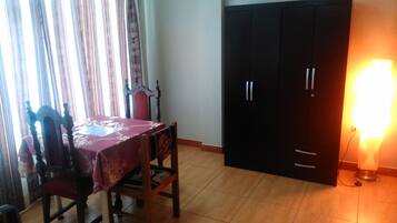 A room with a dining table covered with a red tablecloth, two wooden chairs, a black wardrobe, and a bright lamp on the floor.