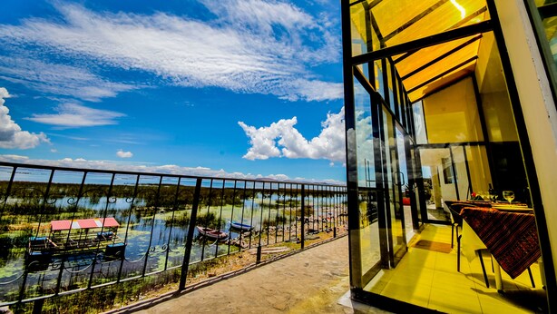 A balcony with a glass railing overlooking a lake with boats, a dining area with a table and chairs, and a yellow interior. - Yacht Lago Titicaca (Puno)