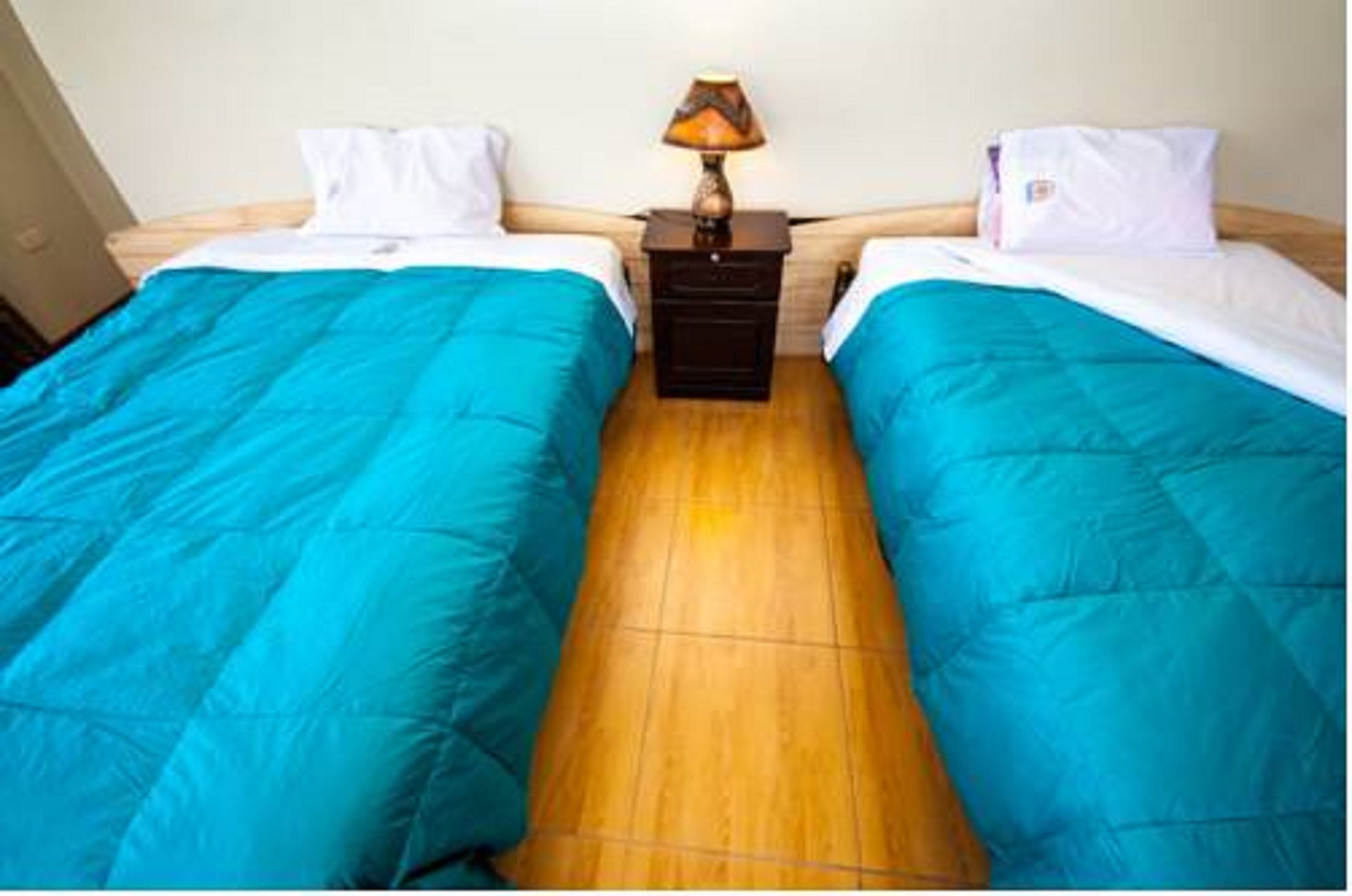 A double room with two beds, each with a blue bedspread and white pillows. There is a nightstand with a lamp between the beds.