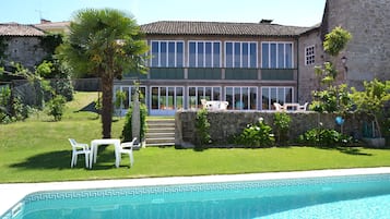 Una casa de dos pisos con piscina, un muro de piedra y un pequeño jardín con sillas y una palmera.