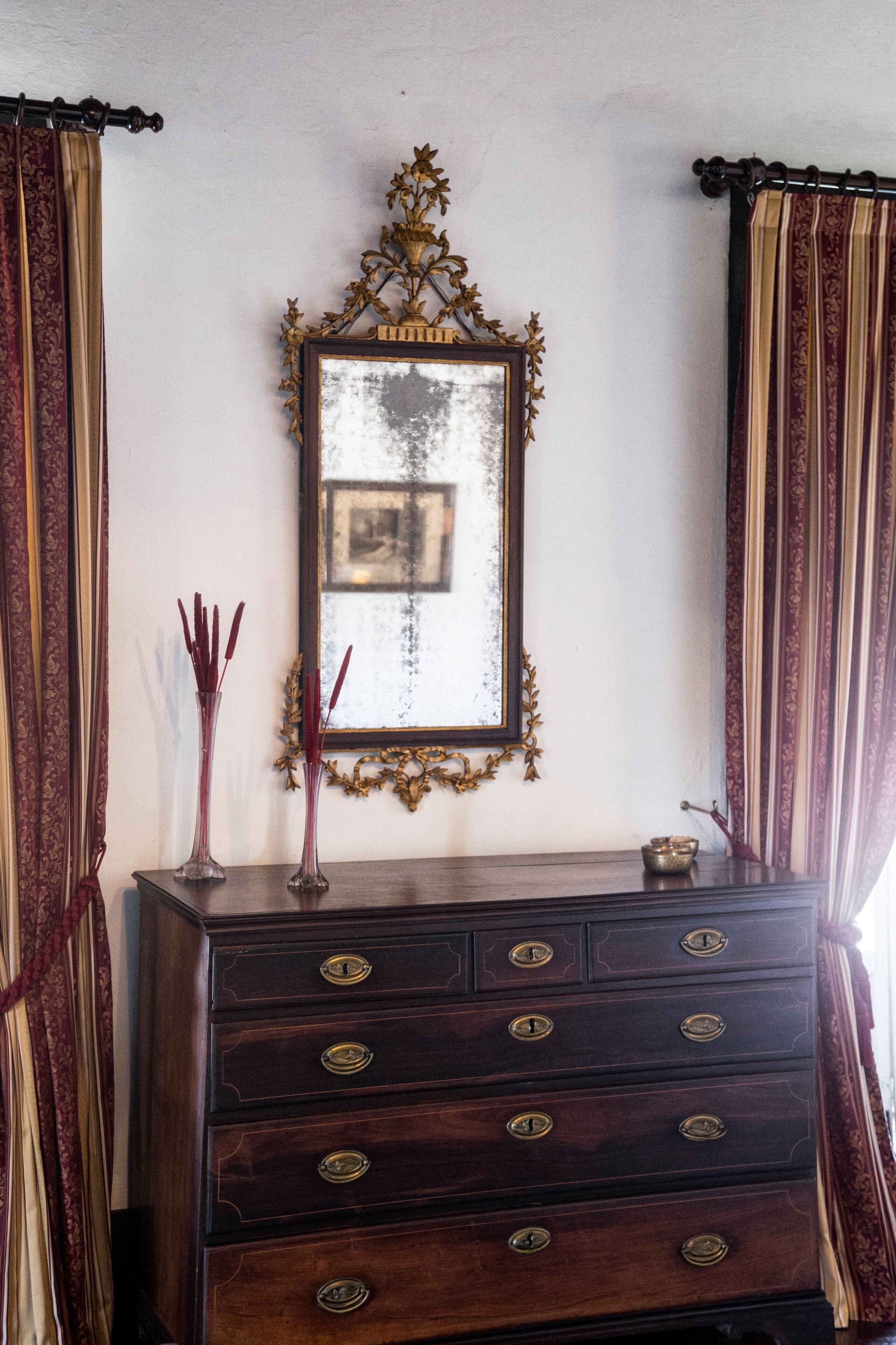 Un aparador de madera con tiradores de latón, un espejo con un marco ornamentado y dos jarrones con flores rojas.
