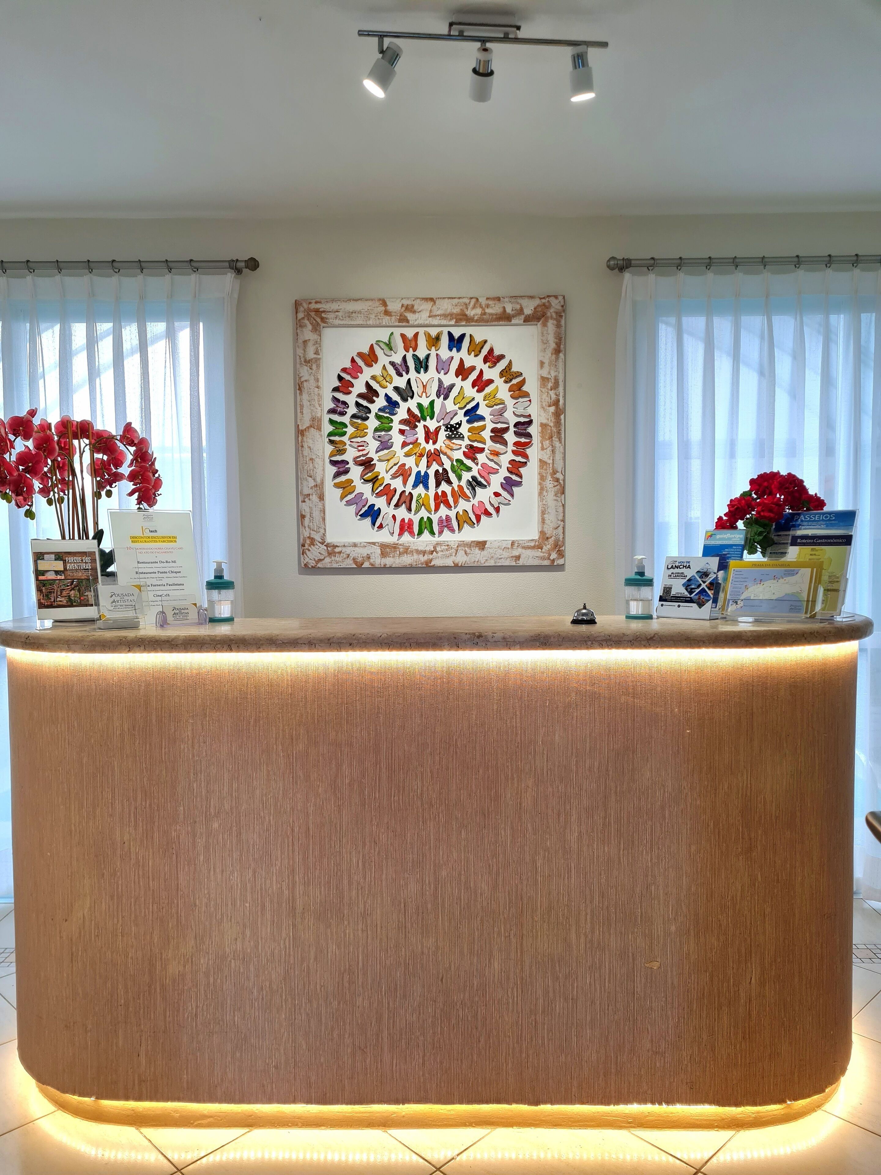 A reception desk with a wooden finish, a framed colorful artwork above it, and a window with sheer curtains on either side.