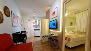 A compact hotel room with a bed, a TV, a desk with a chair, and a kitchenette.