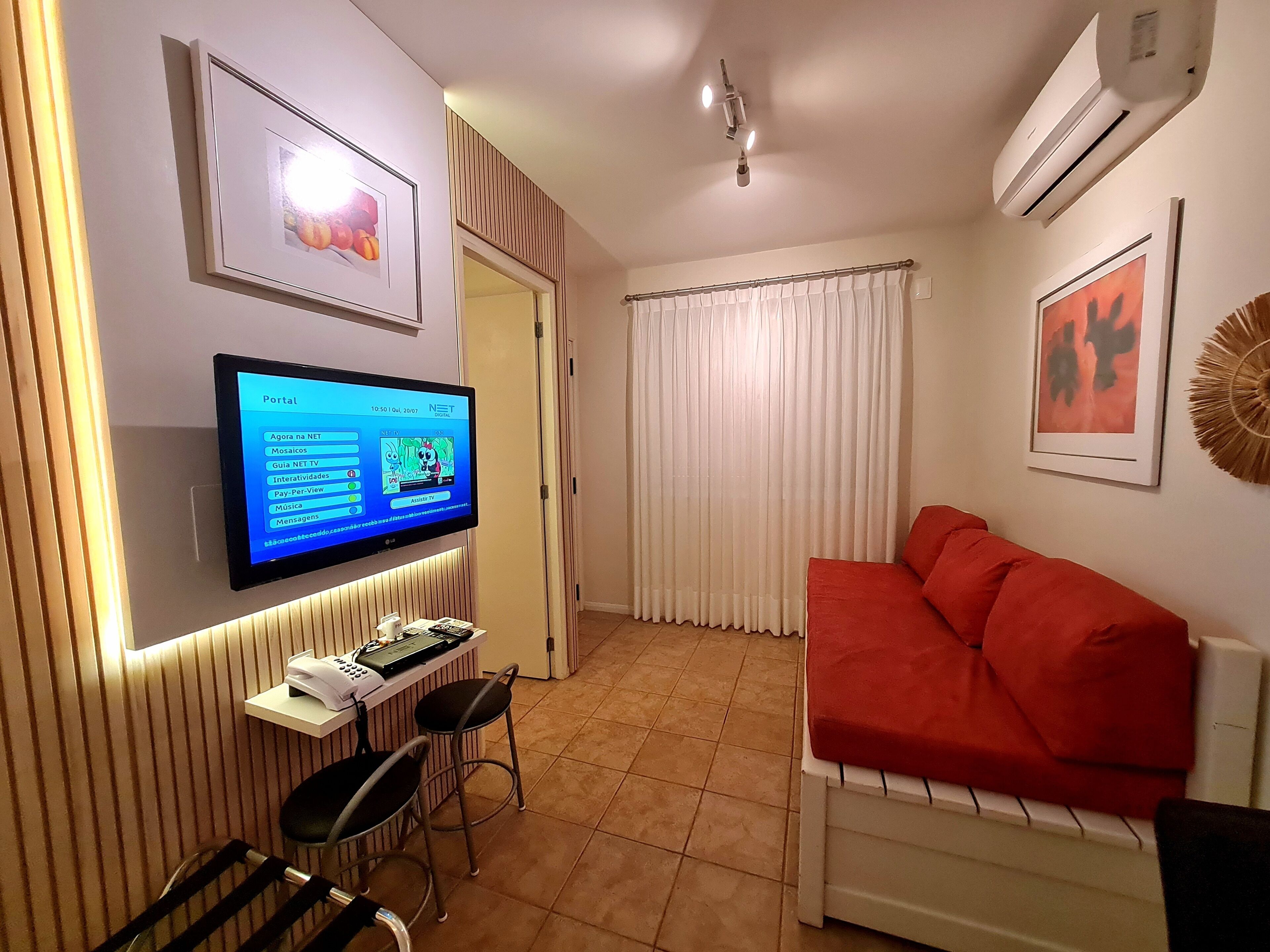 A compact living space with a flat-screen TV mounted on the wall, a red sofa, a small table with a chair, and a window with white curtains.