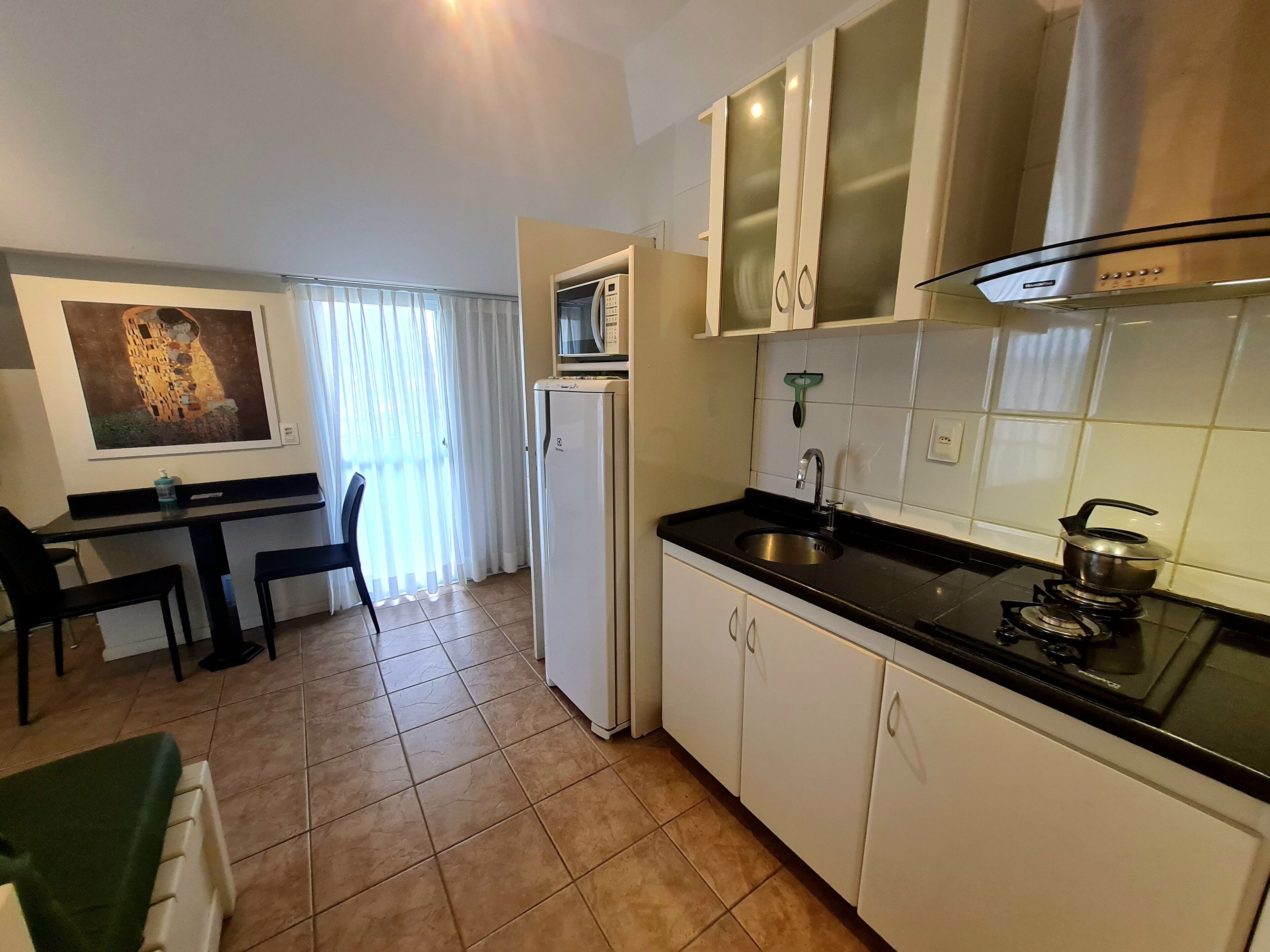 Cozinha compacta com geladeira, micro-ondas, pia e fogão. Há uma área de refeições com mesa e cadeiras.