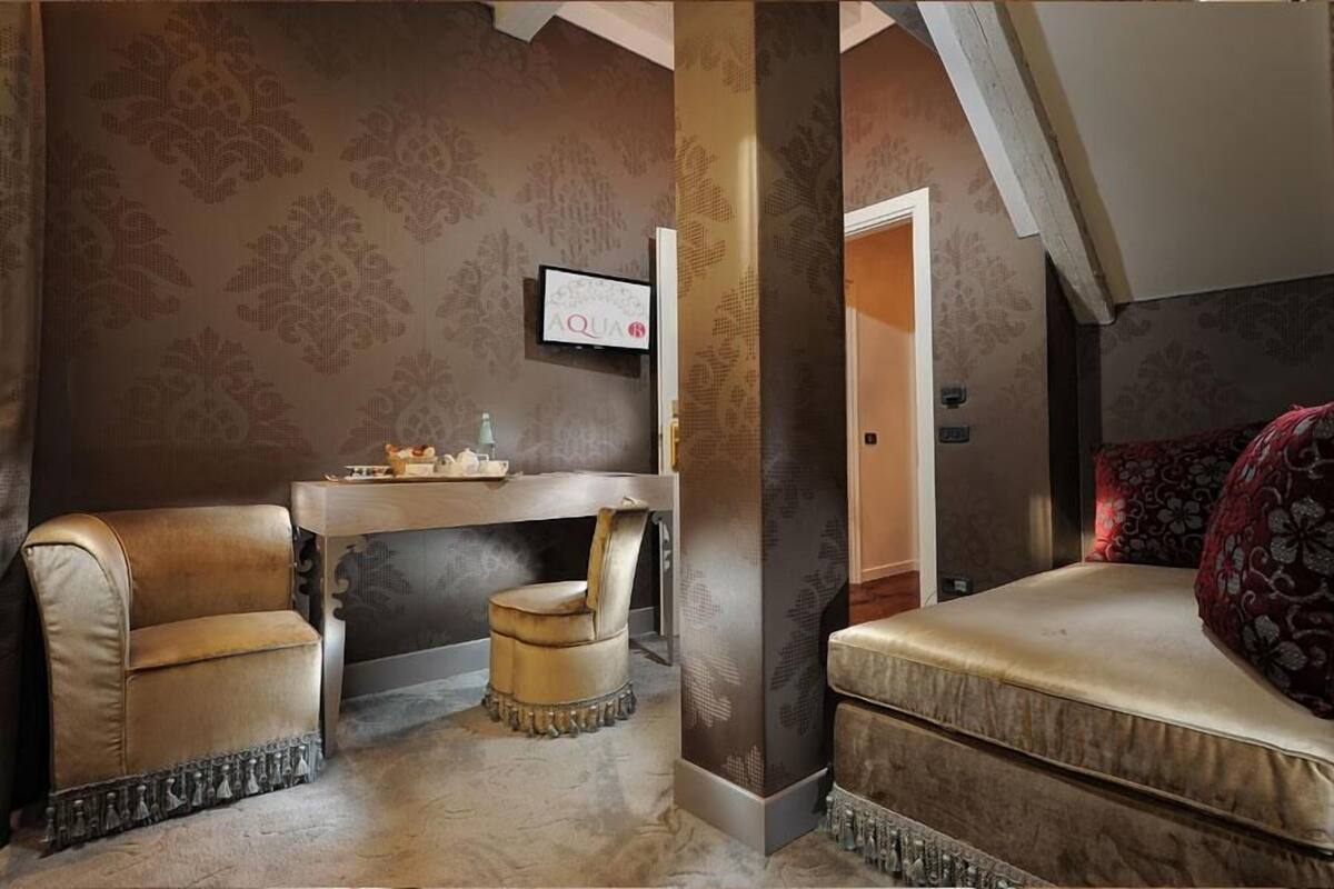 A room with a desk, two chairs, a bed, and a television on the wall.