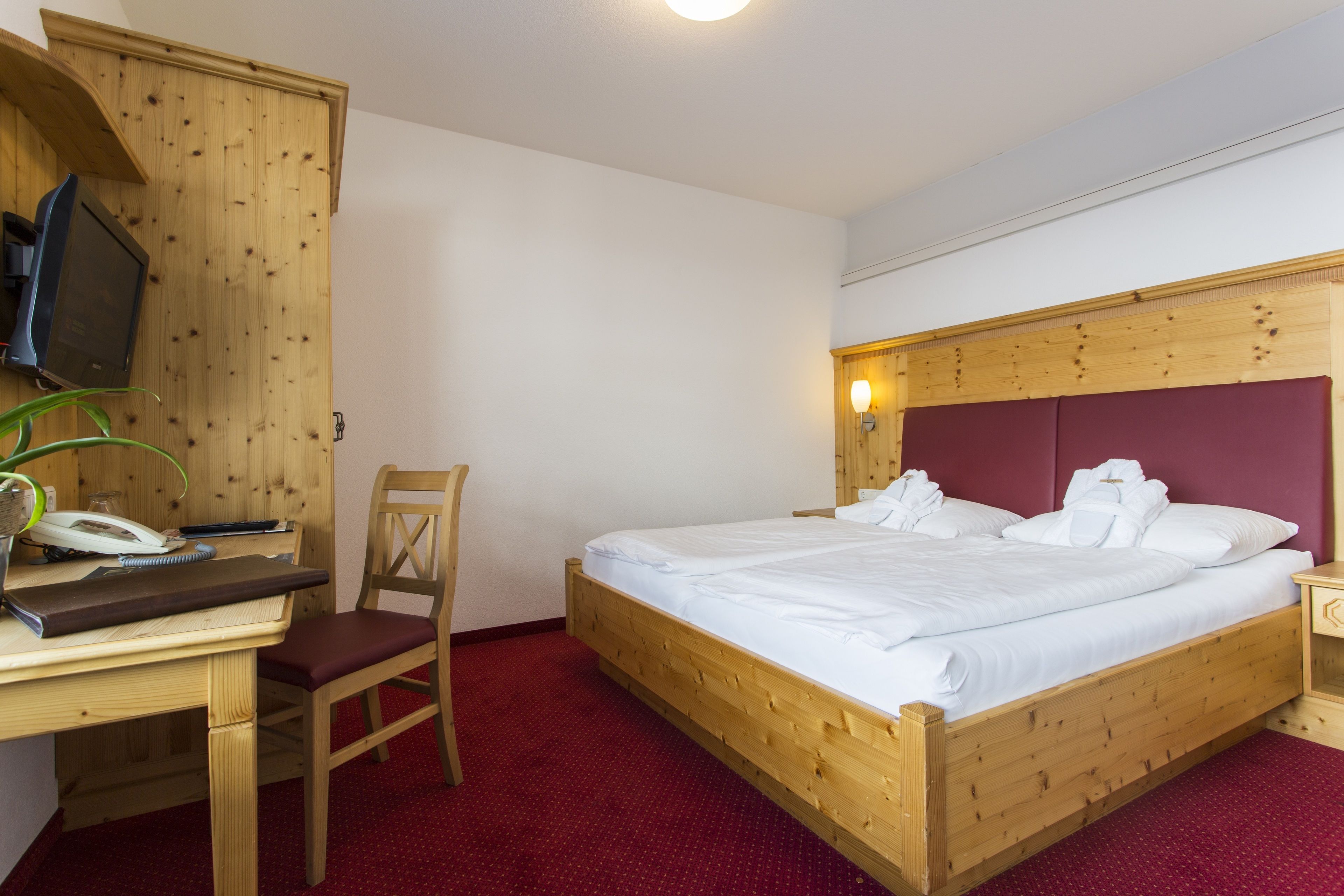 A wooden bed with white bedding, a wooden desk with a telephone, a chair, and a television mounted on a wooden cabinet.