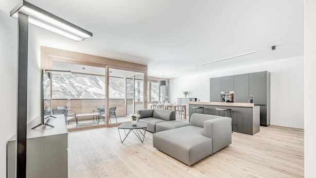 A modern living room with a grey sectional sofa, a coffee table, and a dining area with a table and chairs. There is a kitchen area with grey cabinets and a countertop. - all inclusive Hotel Lohmann (Soelden)