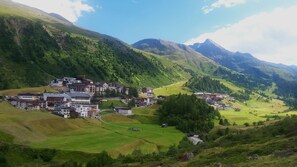 A mountainous village with buildings clustered on a hillside, surrounded by lush greenery and mountains.