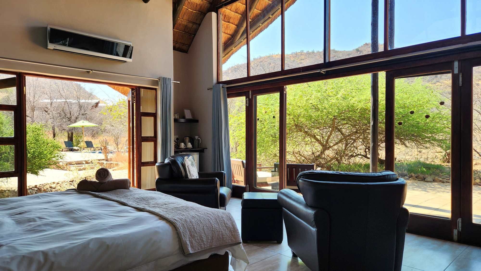 A bedroom with a large bed, a sofa, and a chair, with a view of a garden and trees through large windows.
