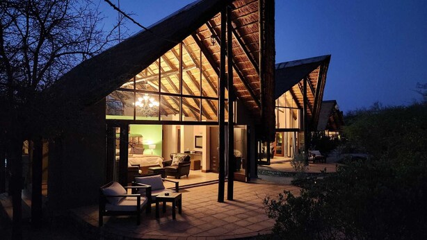 A modern house with a large glass facade, a dining area with outdoor seating, and a well-lit interior.