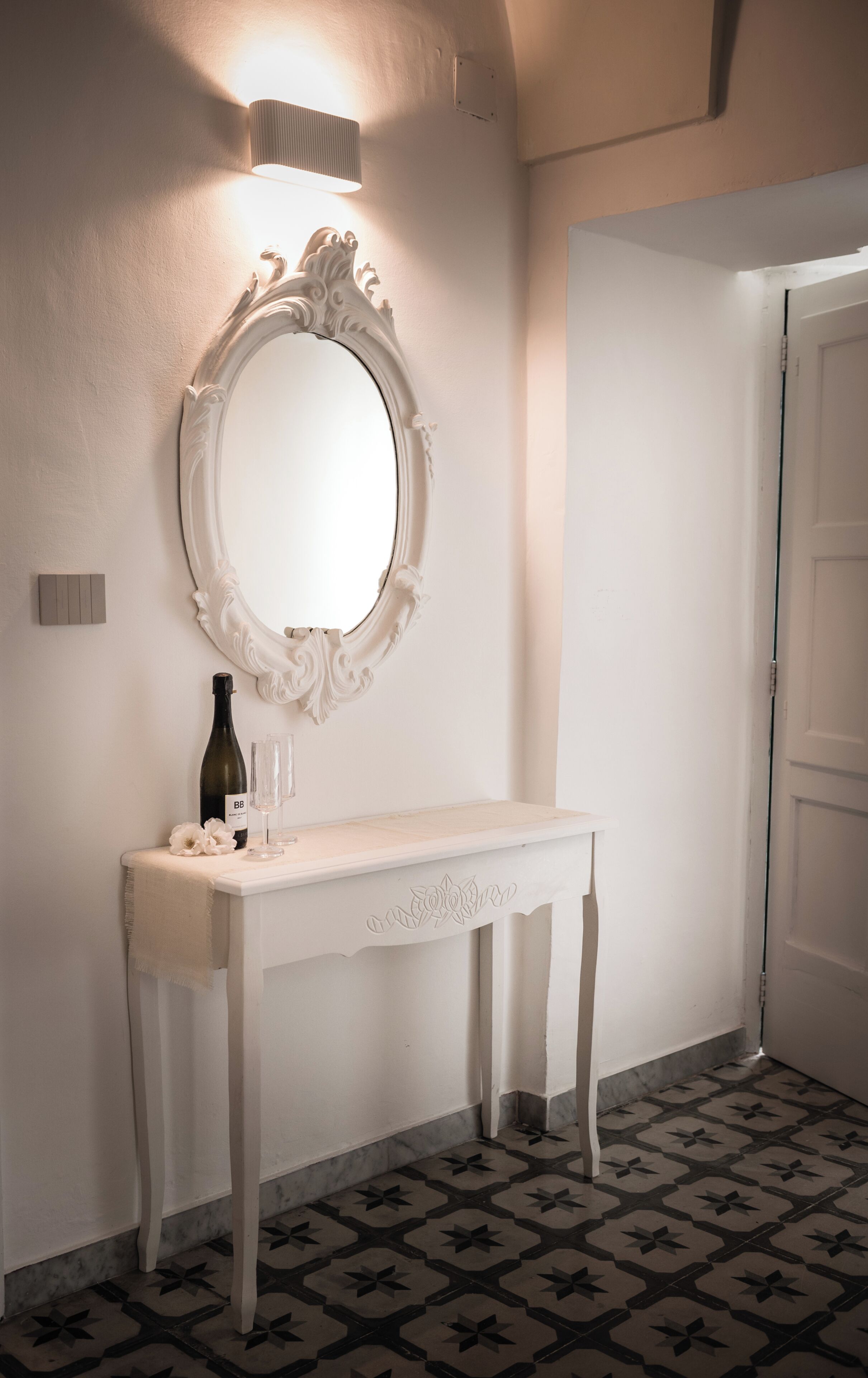 A bathroom with a white vanity, a mirror with an ornate frame, a bottle of wine, and two glasses.
