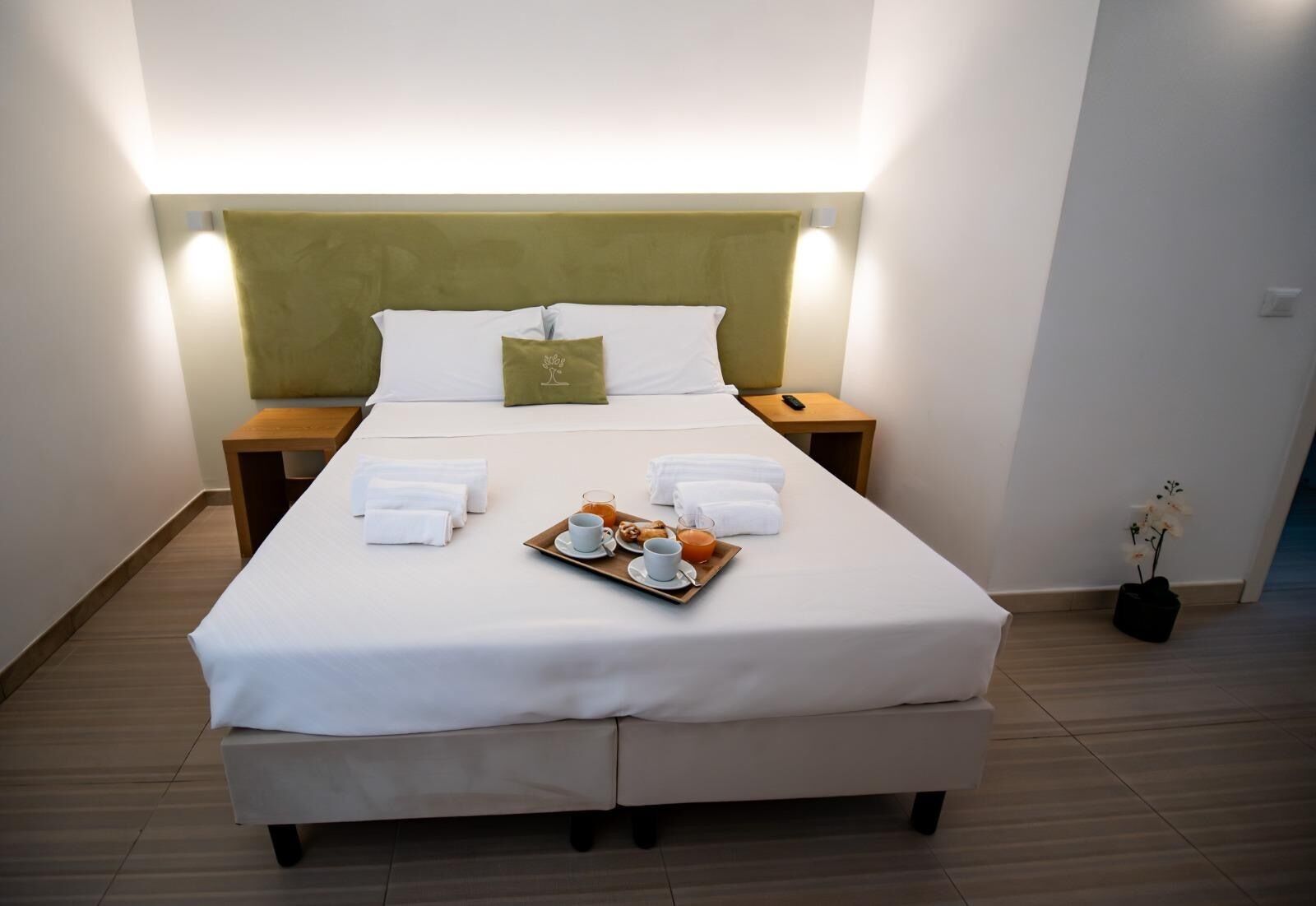 Un lit bien fait, avec un plateau contenant des articles pour le petit-déjeuner, une table de chevet avec une télécommande et une petite plante en pot.
