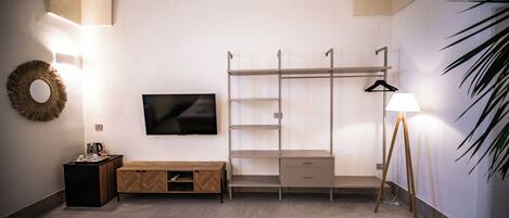 Habitación de hotel con cama, televisor, banca de madera, armario, perchero, lámpara y planta.