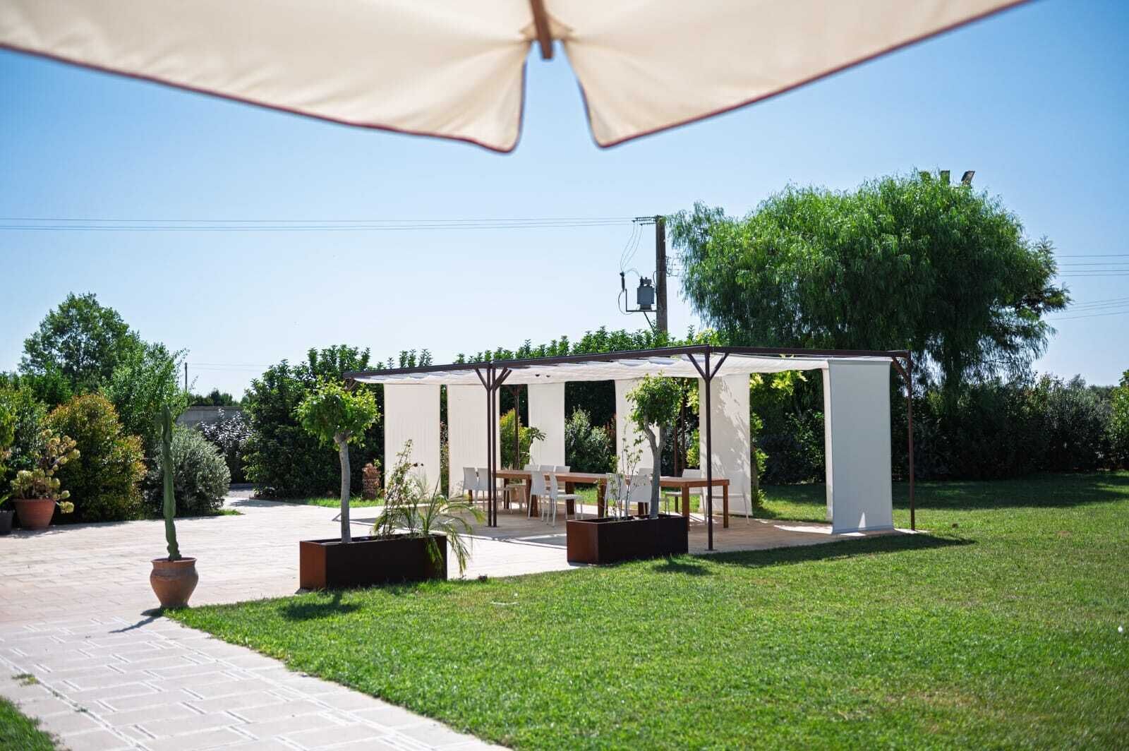 A covered outdoor patio with a table and chairs, surrounded by greenery and a clear sky.