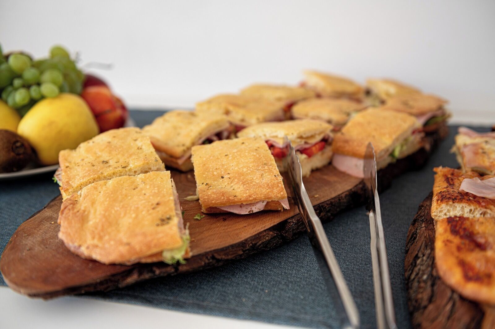 Een houten plank met verschillende sandwiches, gesneden in vierkanten, vergezeld van een bord fruit en een tang.