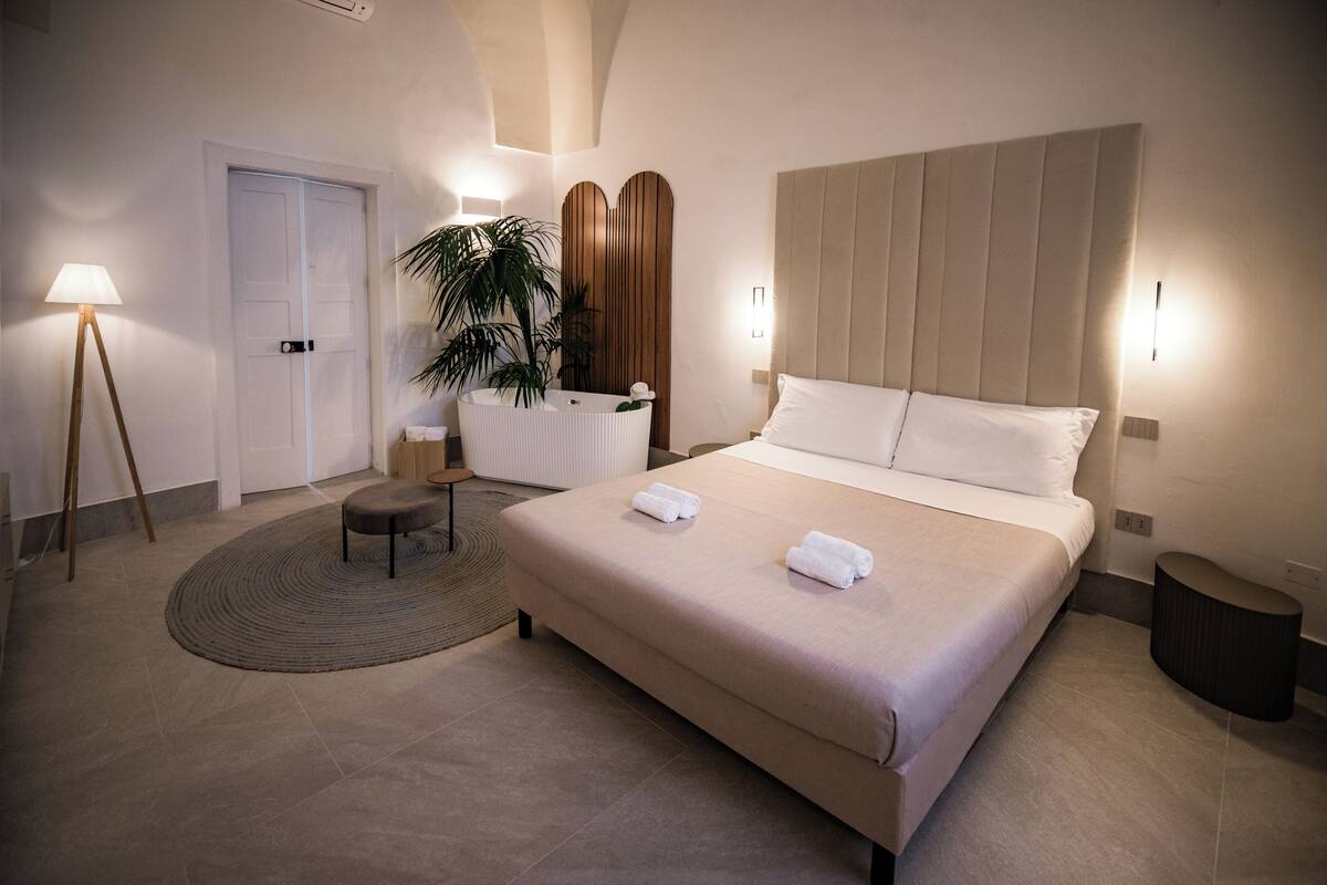 A bedroom with a large bed, bedside tables, a round rug, a plant, and a door.