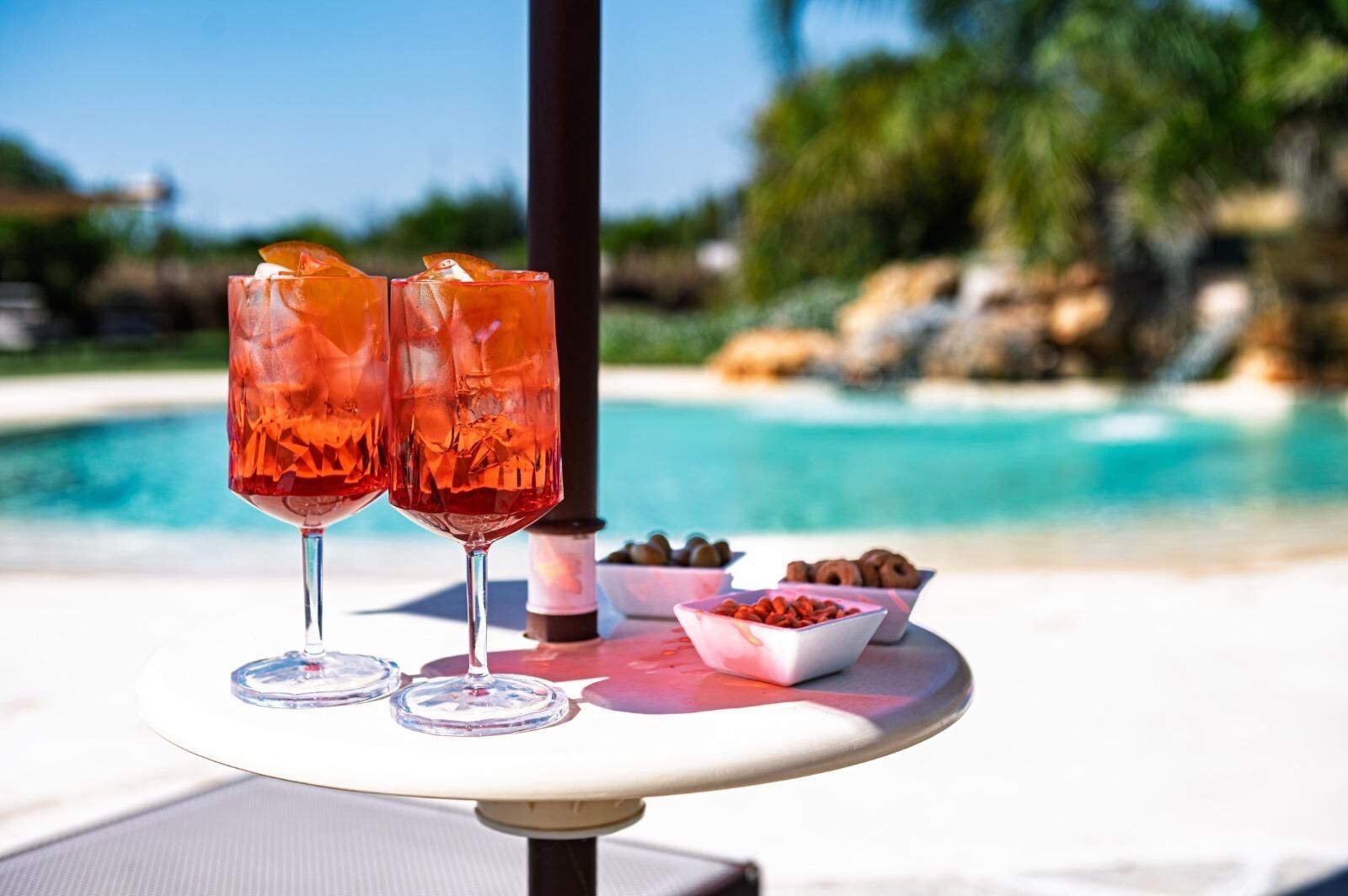 Dos vasos de bebida roja sobre una mesa junto a una piscina, con aperitivos.