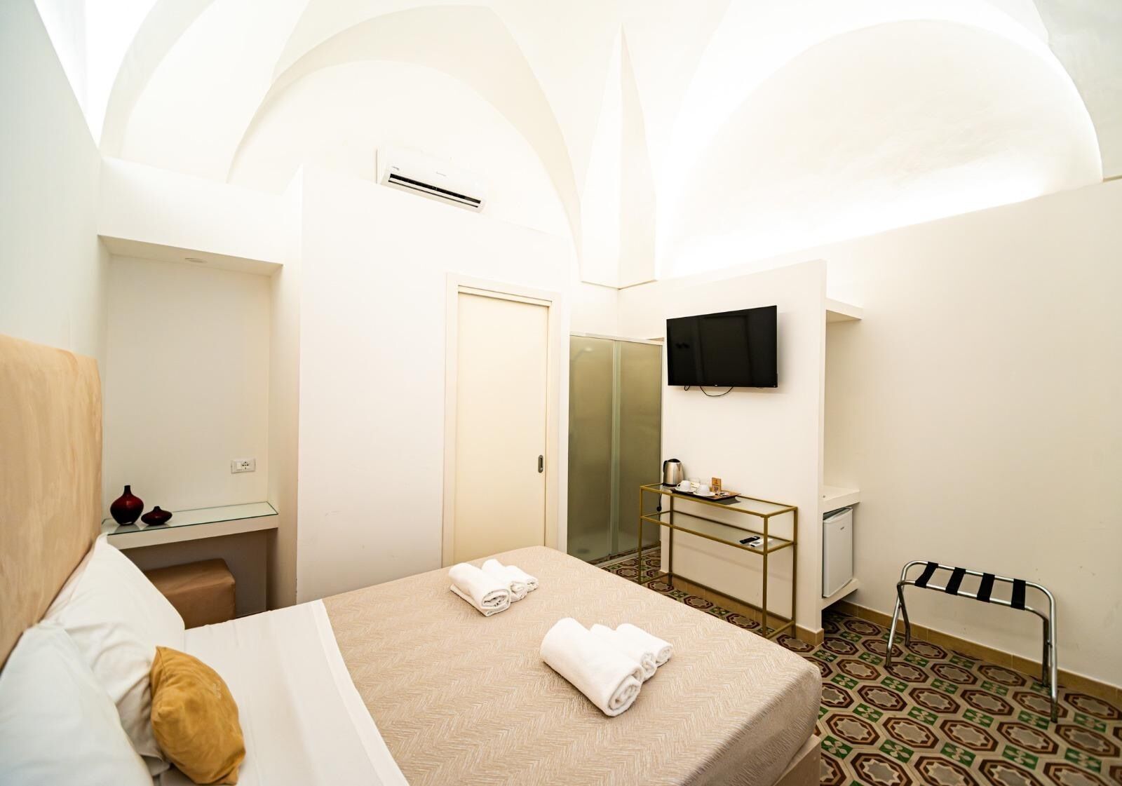 A hotel room with a bed, a TV, a mirror, a small table with a kettle, and a shower.