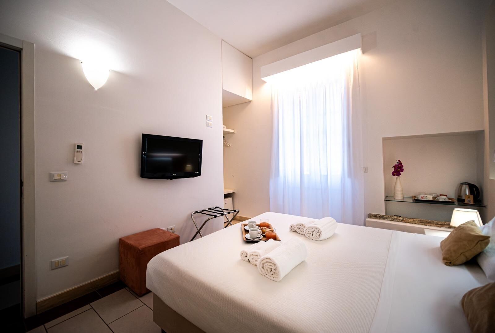 A hotel room with a bed, a TV, a window with curtains, a small table with towels and a tray of food, and a shelf with toiletries.