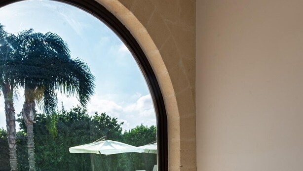 A room with a view of a pool, a table with chairs, and a large arched window. - Tenuta San Nicola (Novoli)