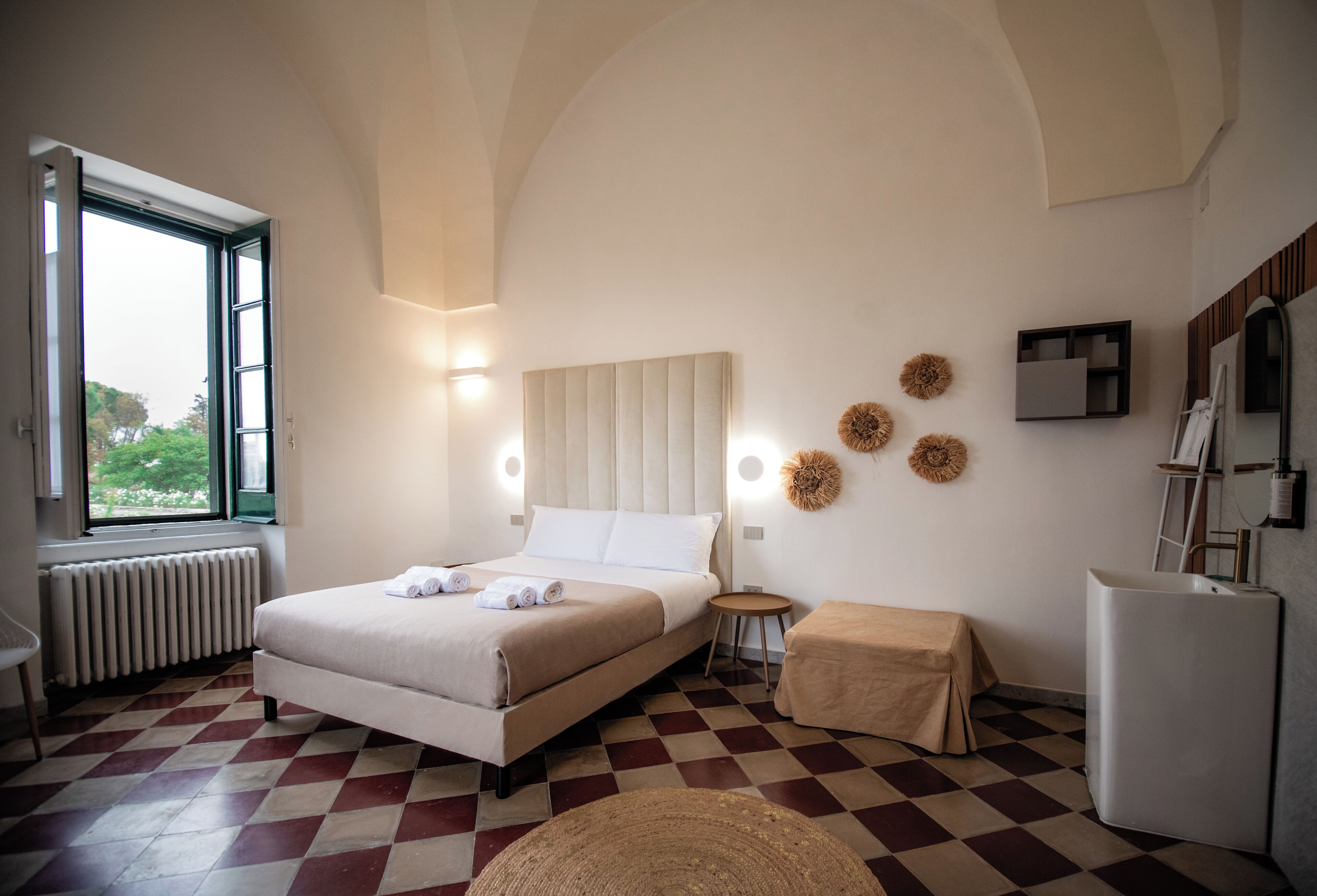 A hotel room with a checkered floor, a bed with a headboard, a small round table, a bedside table, a radiator, and a window with a view of greenery.