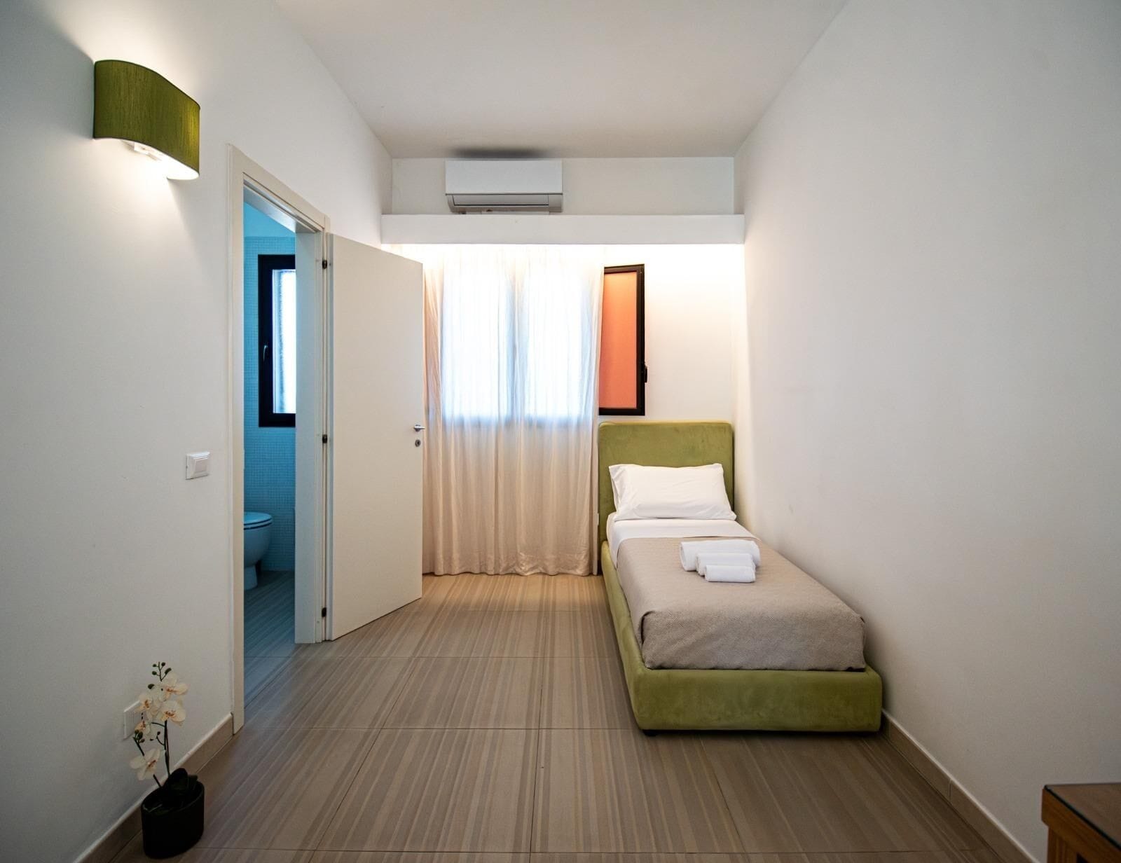 A small, well-lit bedroom with a bed, a window with curtains, a wall-mounted air conditioner, and a door leading to a bathroom.