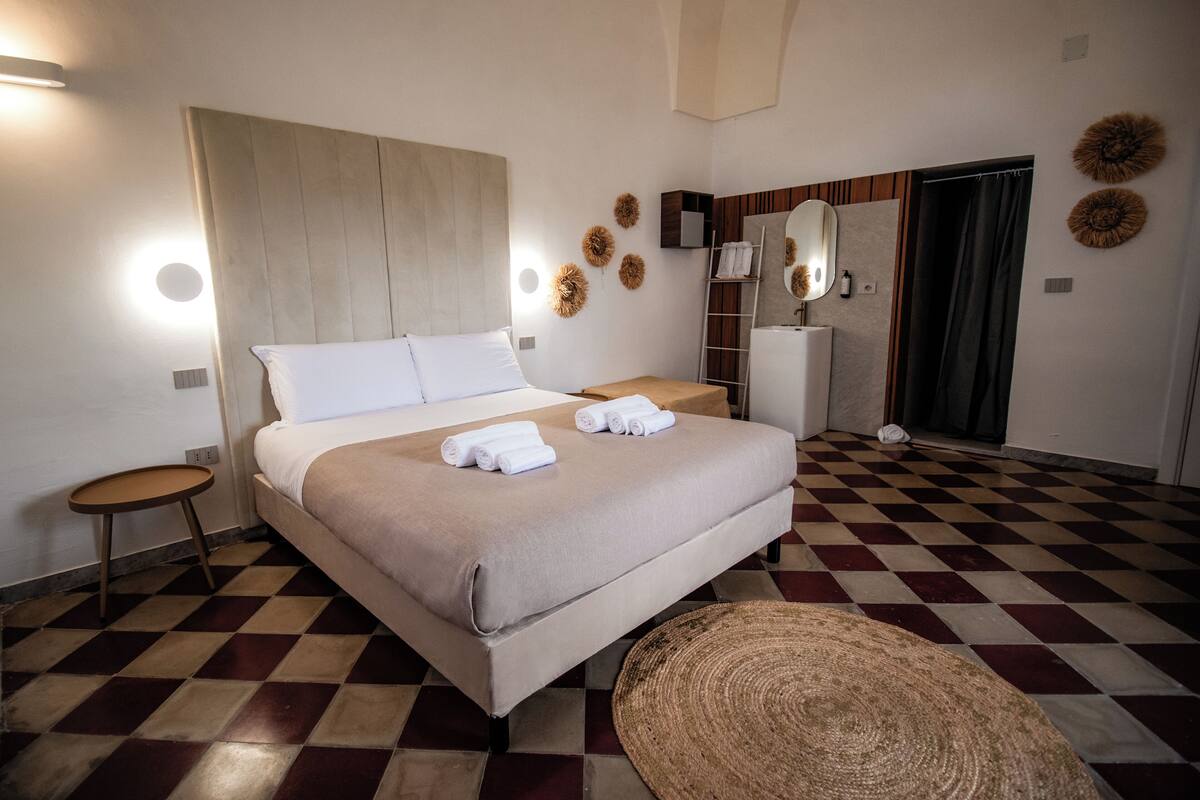 A hotel room with a checkered floor, a bed with white linens, a round woven rug, and a mirror on the wall.