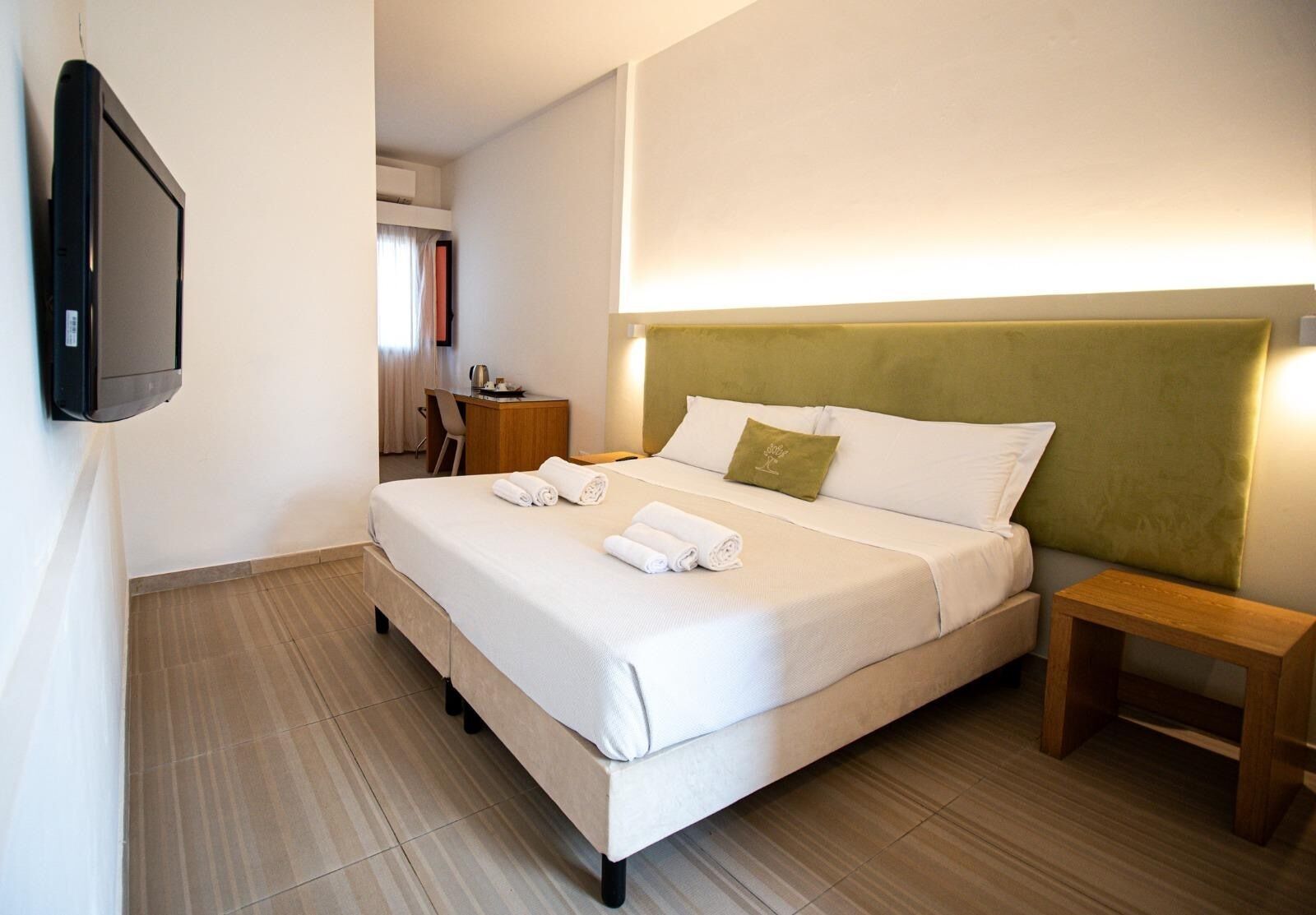 Ein Hotelzimmer mit Bett, Nachttisch, einem an der Wand befestigten Fernseher und einem Schreibtisch mit Stuhl.