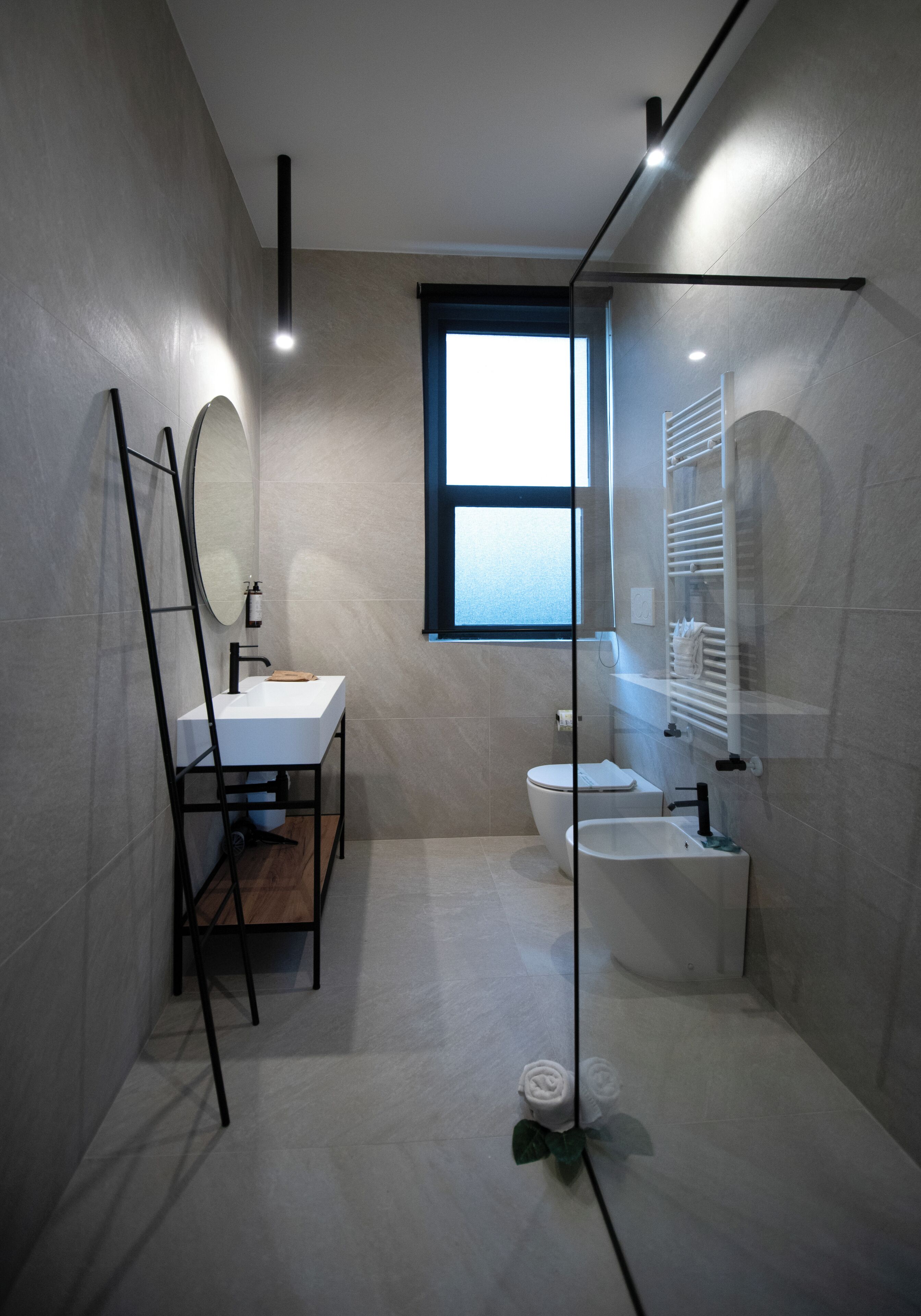 A modern bathroom with a white sink, toilet, and bidet, a mirror, and a window.