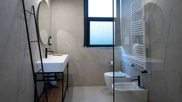 A modern bathroom with a white sink, toilet, and bidet, a mirror, and a window.