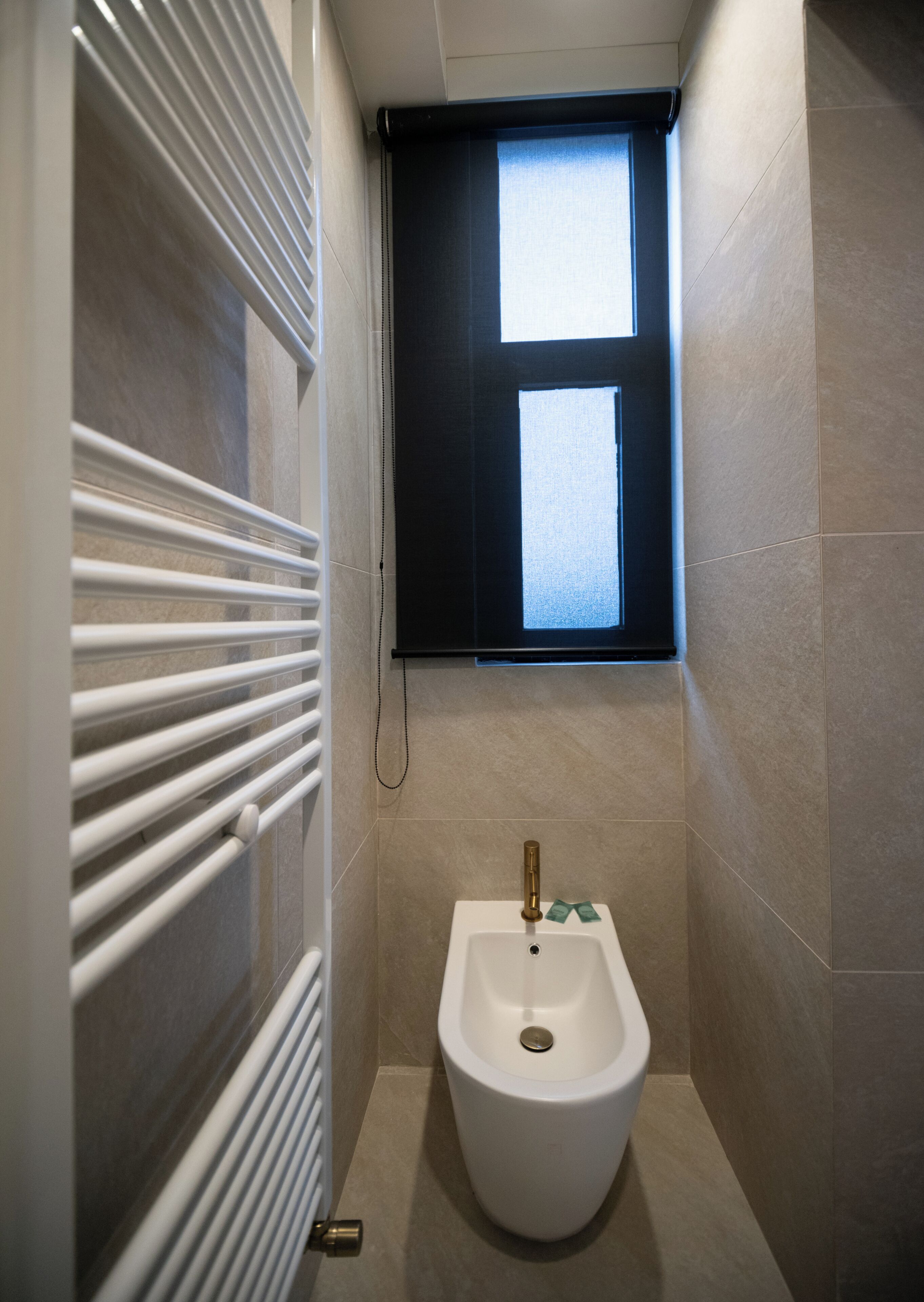 A small, modern bathroom with a bidet, a window with blinds, and a radiator.