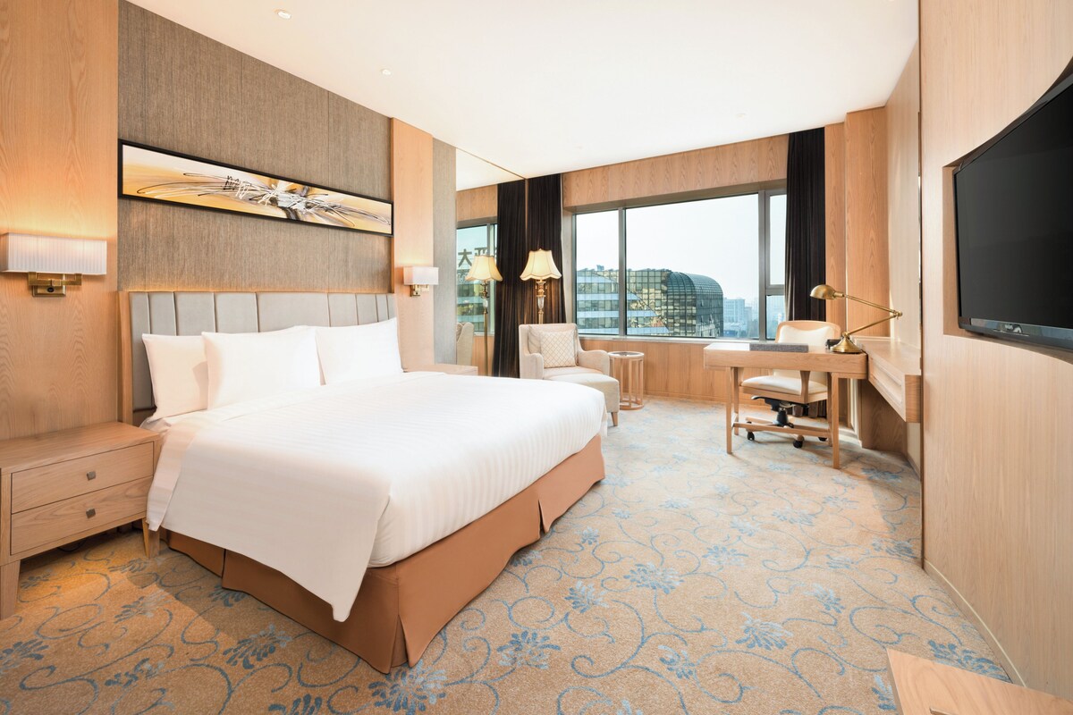 A hotel room with a large bed, a desk, a chair, a TV, and a window with a city view.