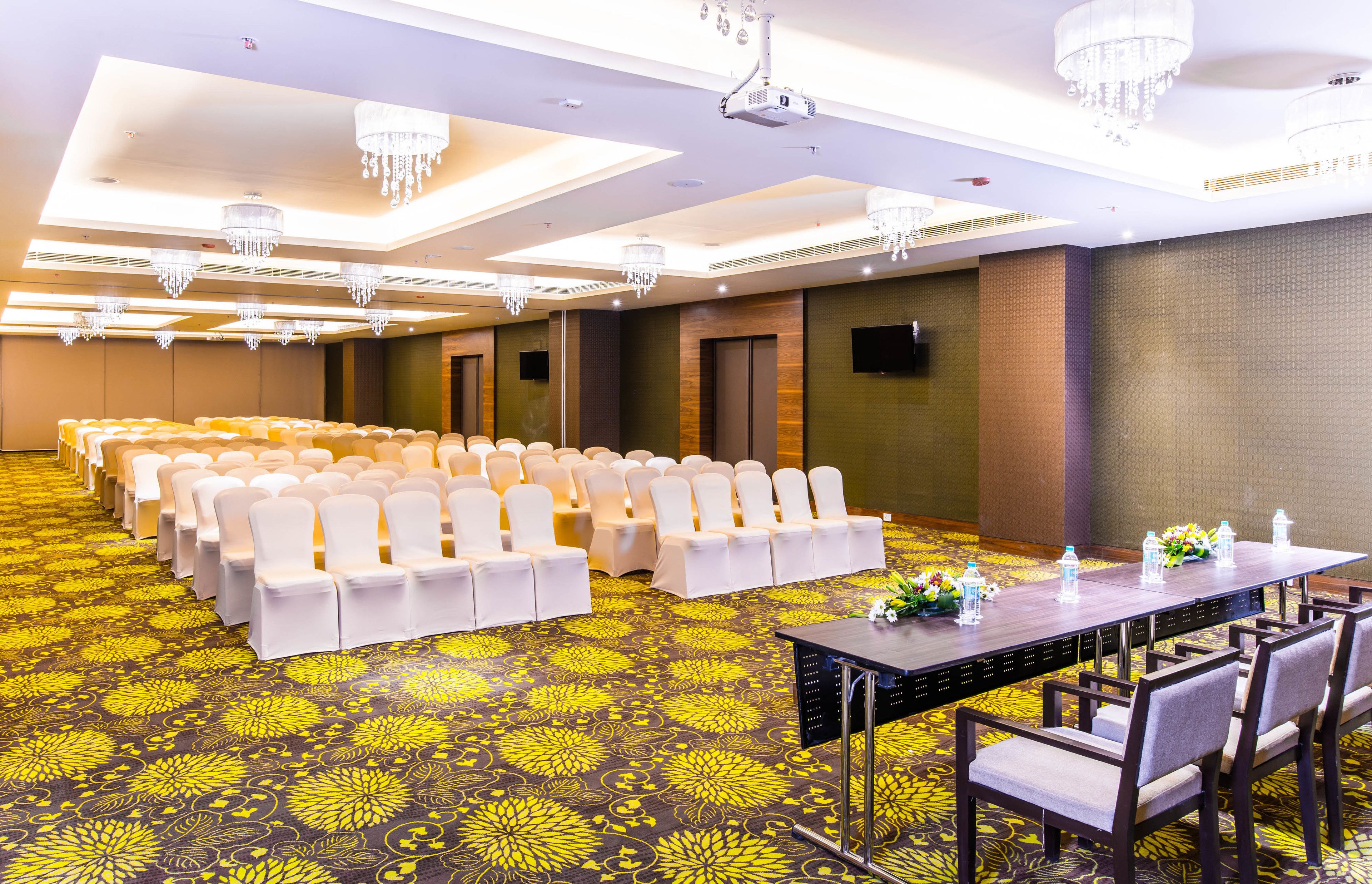 Un salón de conferencias con filas de sillas blancas, una larga mesa con botellas de agua y una alfombra con estampado floral amarillo.