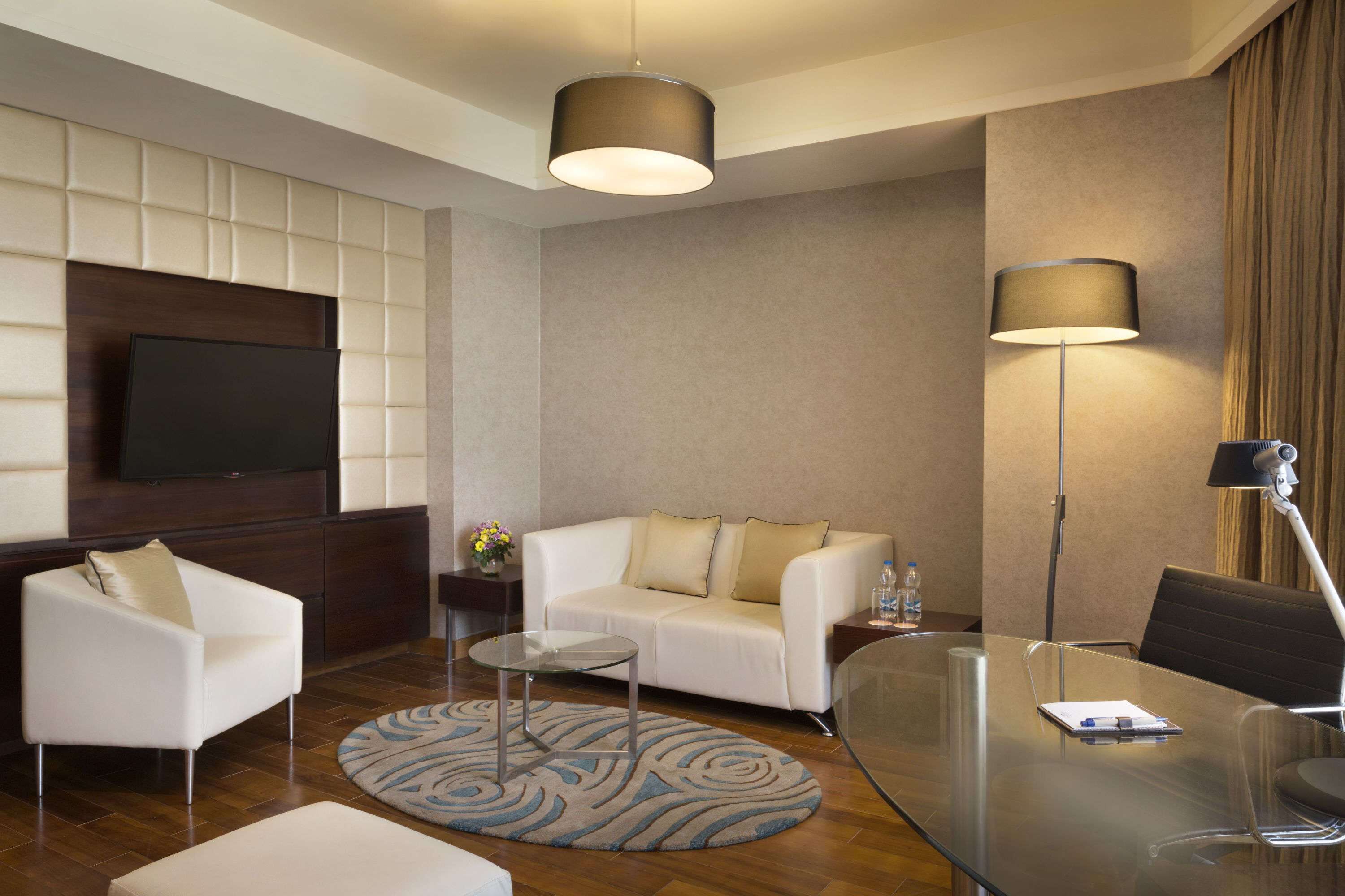 A modern hotel room with a sofa, armchair, glass table, and a flat-screen TV mounted on the wall.