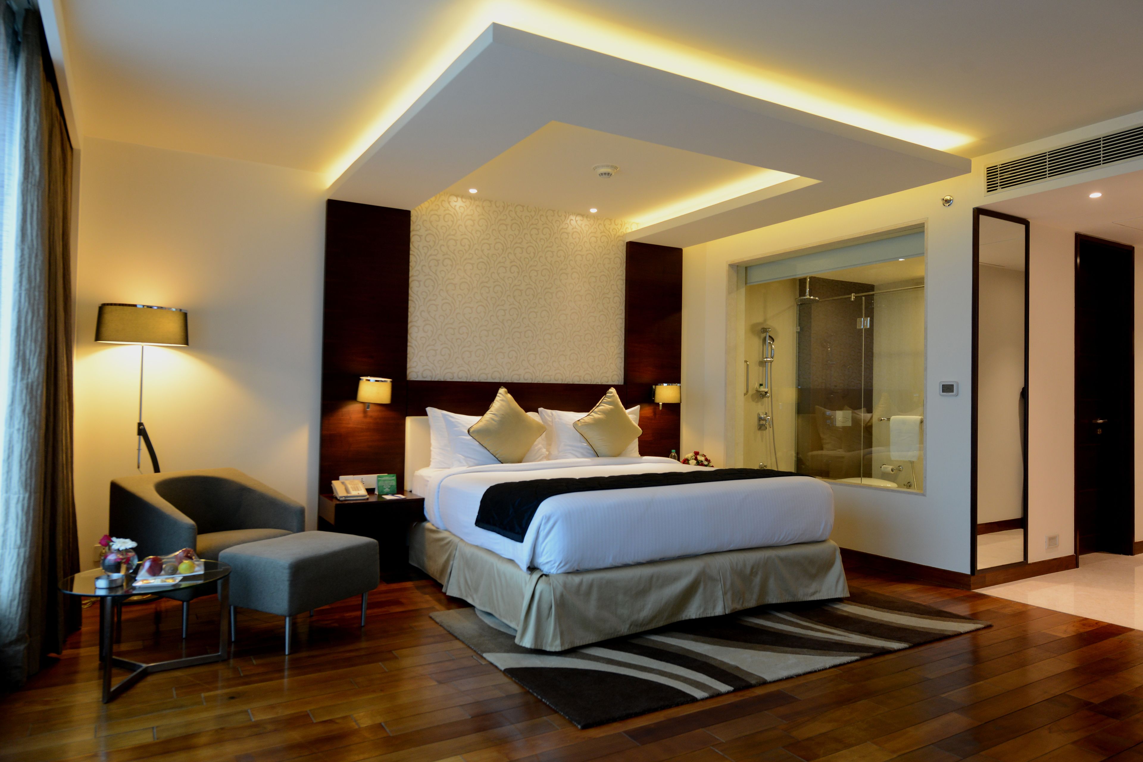 A modern hotel room with a large bed, a sitting area, a desk, and a bathroom with a glass shower.