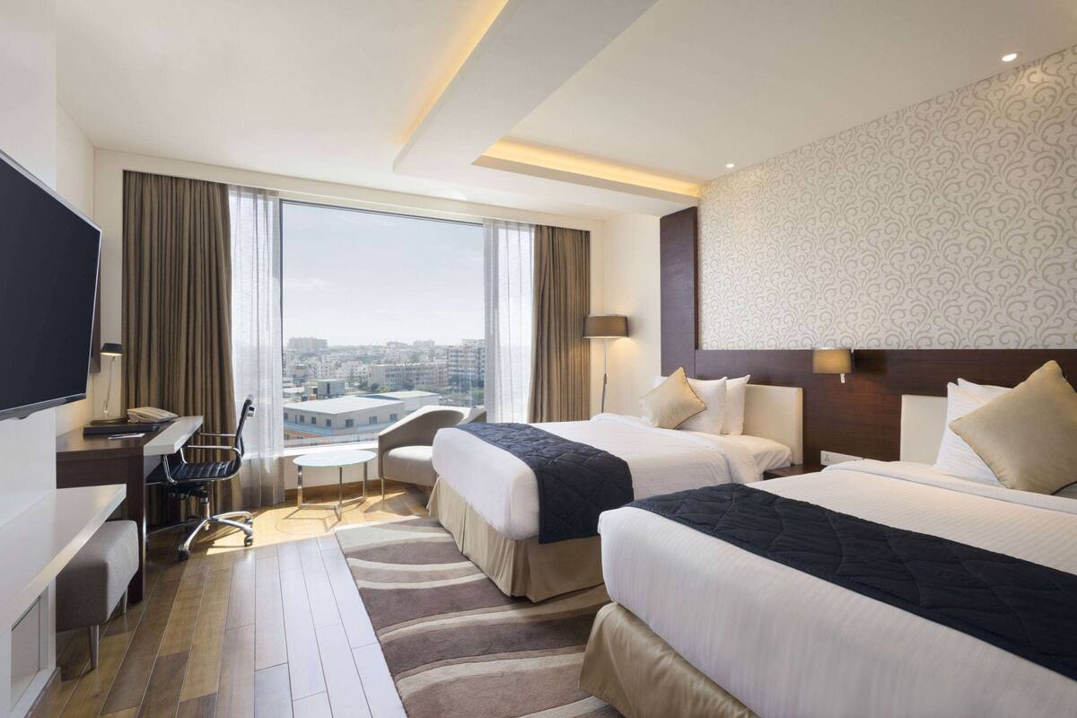 Une chambre d’hôtel avec deux lits, un bureau, une chaise et une grande fenêtre donnant sur la ville.