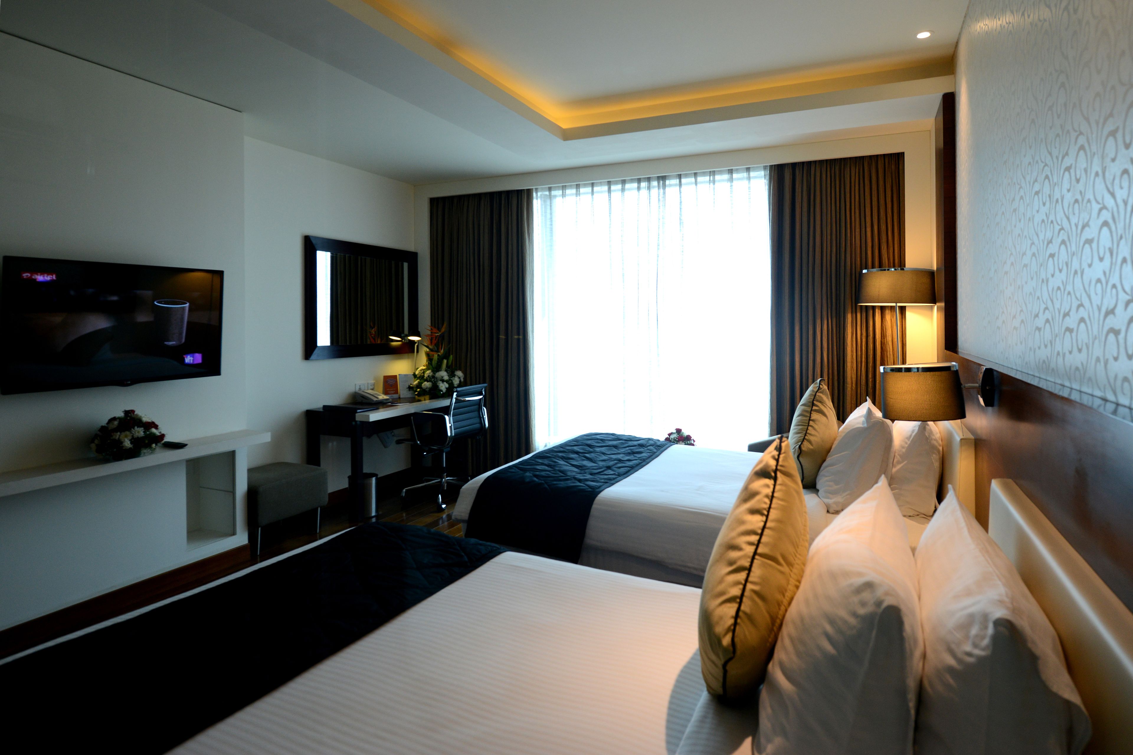 A hotel room with two beds, a desk, a TV, and a large window with curtains.
